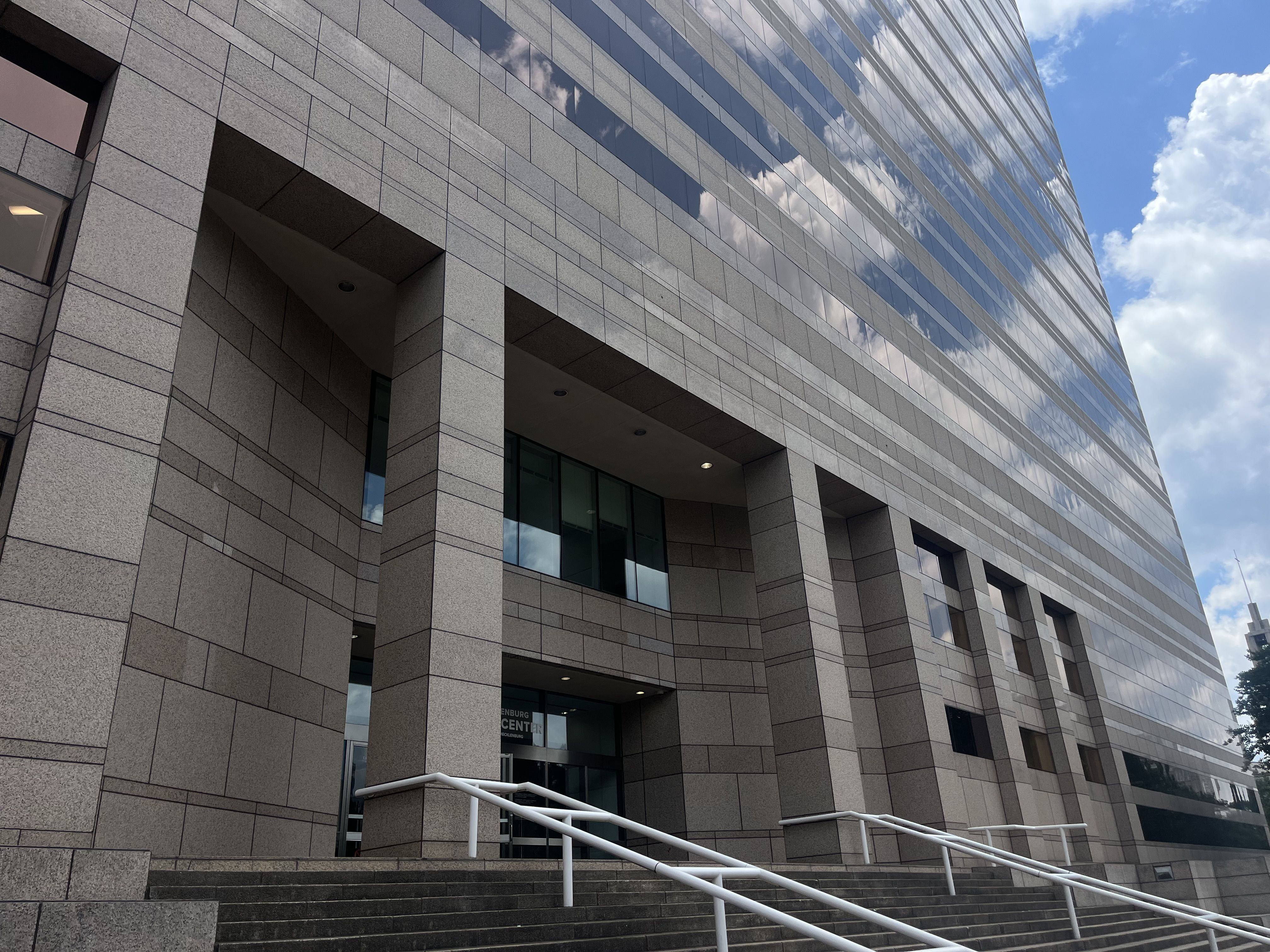 Low-angle view of a modern grey stone building with reflective glass windows along the right, mirroring blue sky and clouds; stairs and white railing in the foreground.