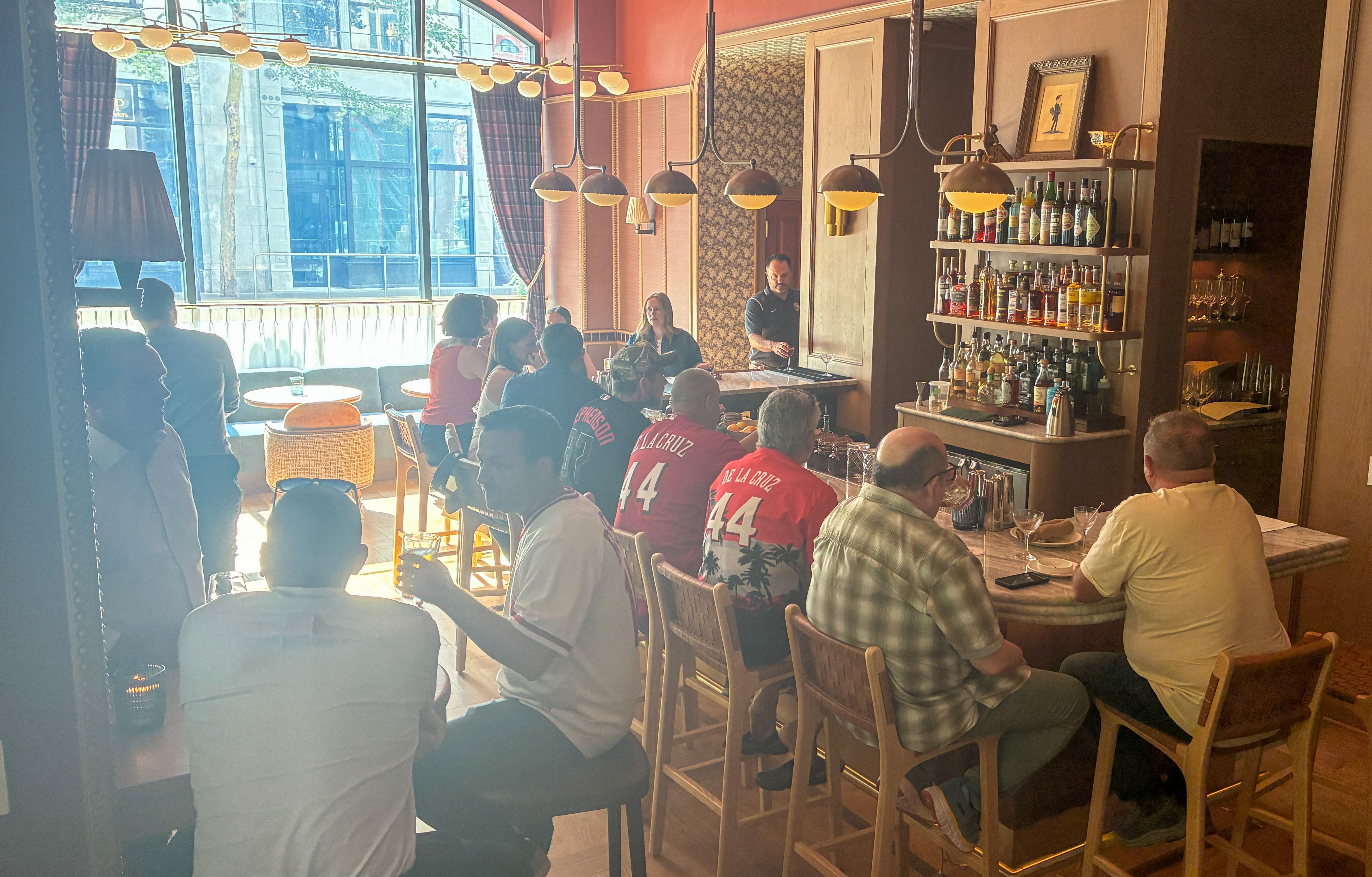 Busy restaurant bar with patrons at tables, many wearing red jerseys. A bartender pours drinks behind a liquor shelf under warm pendant lights beside large windows.