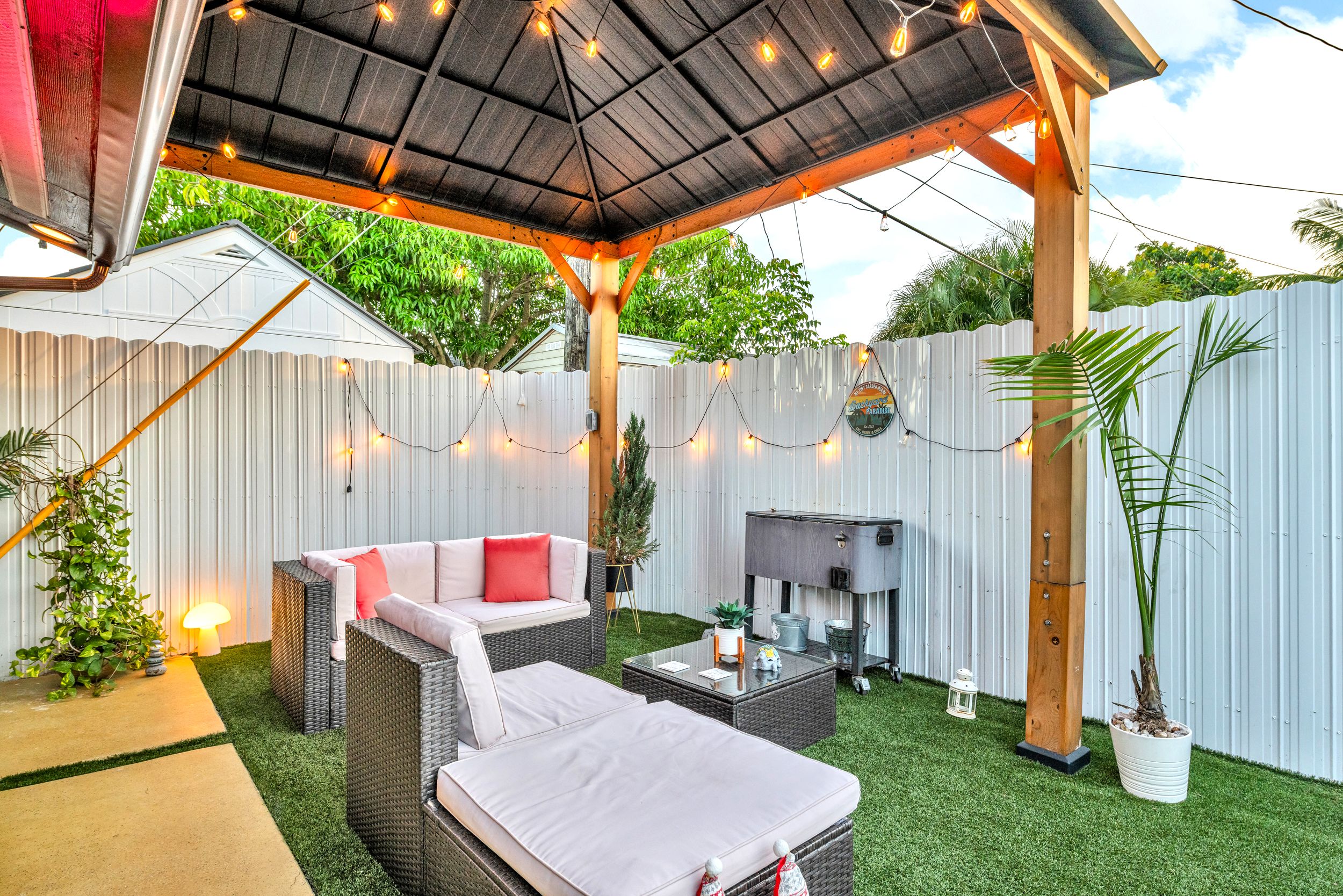 Outdoor patio with black roof, string lights, gray wicker furniture with white cushions and red pillows, green artificial grass, potted plants, and a gray cooler against a white fence.
