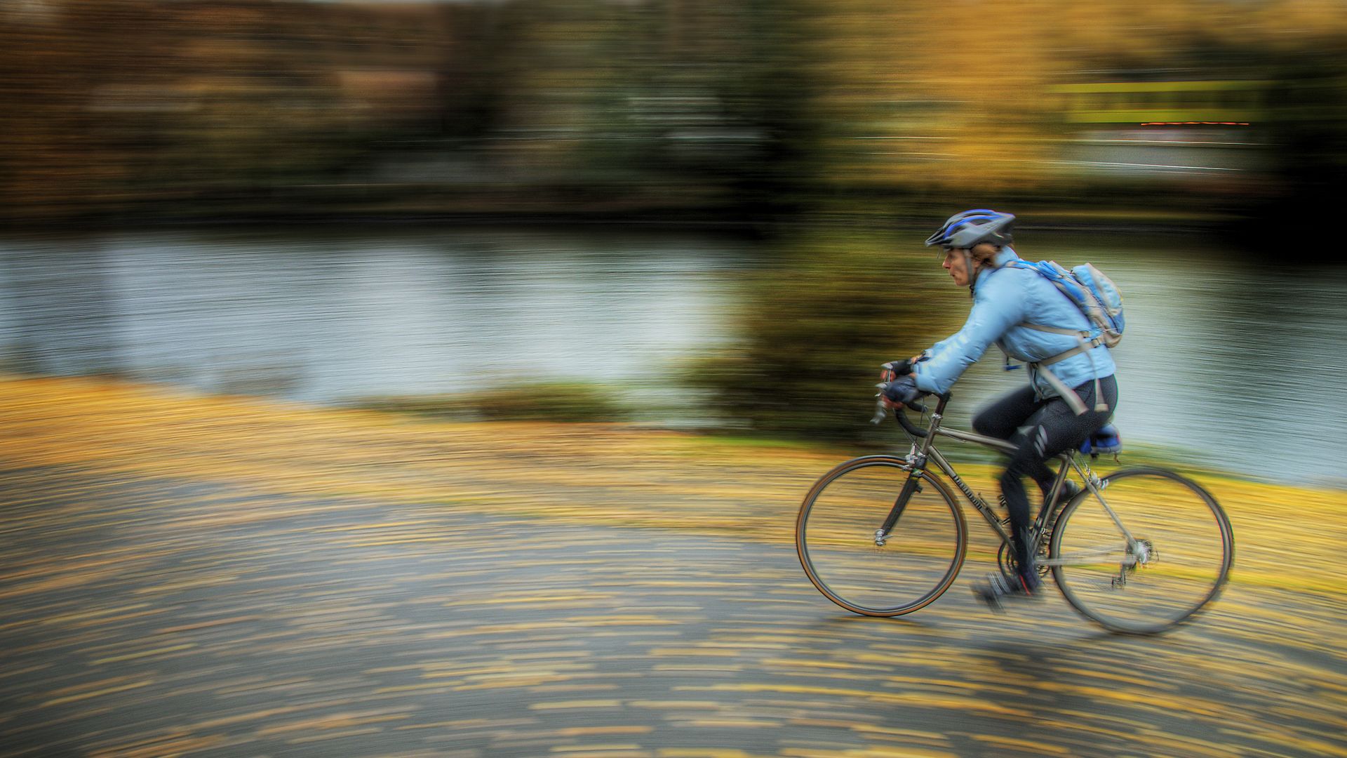 A person in a blue jacket rides by in a blur along a trail strewn with yellow leaves. 
