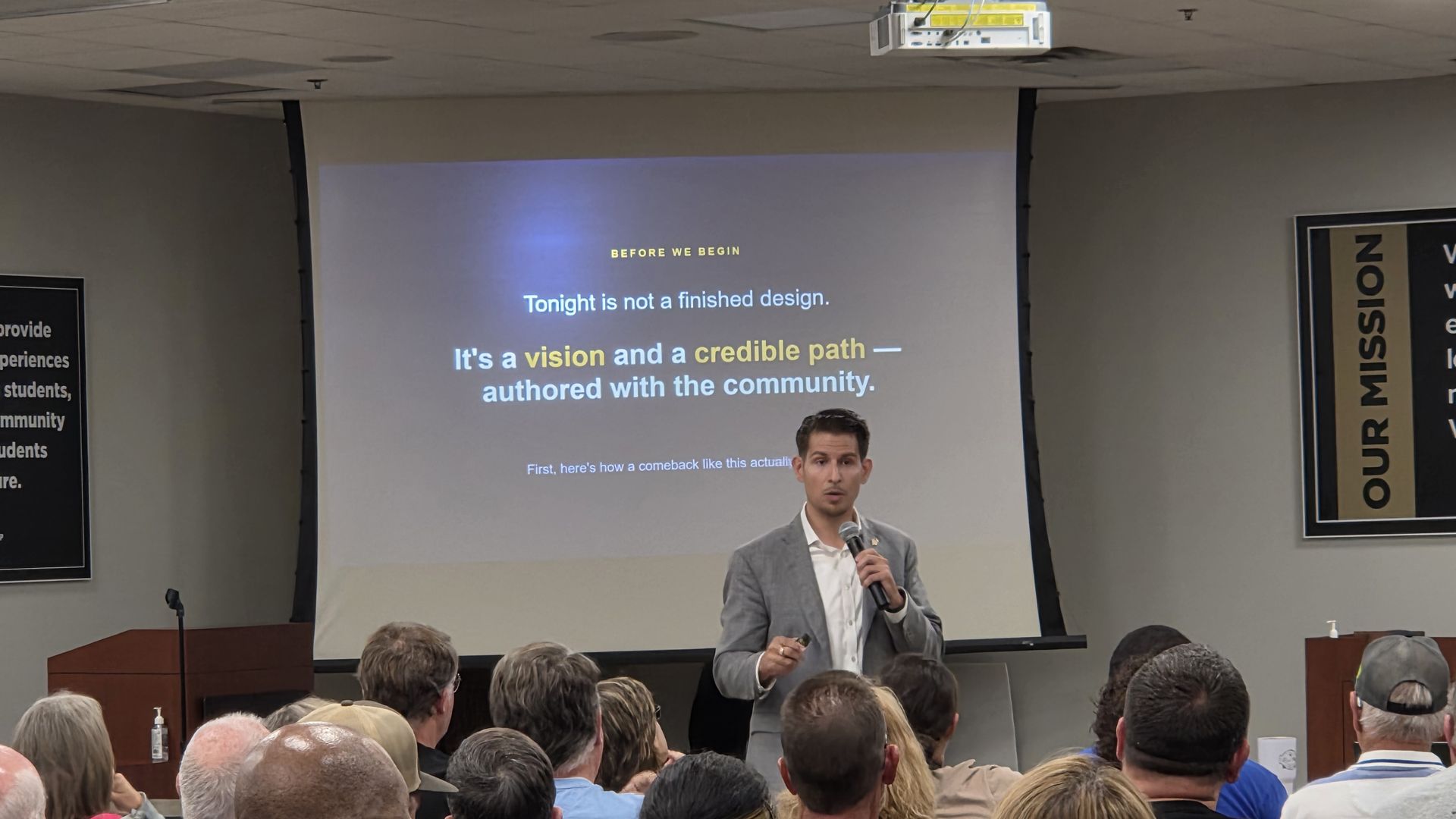 Presenter in a gray suit with a microphone at a podium, showing a slide titled "Washington Square" to a packed audience; wall banners read "OUR MISSION" and "VISION".