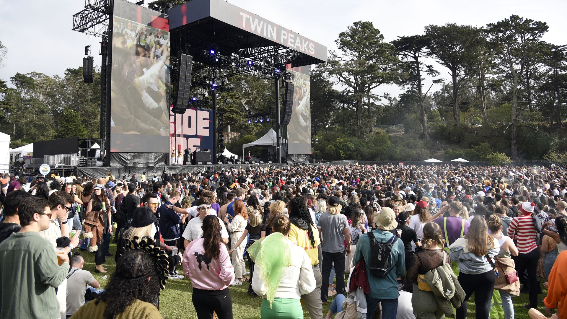 Large crowd of people gathered on grass in front of the Twin Peaks stage at an outdoor concert, surrounded by trees under a bright sky.