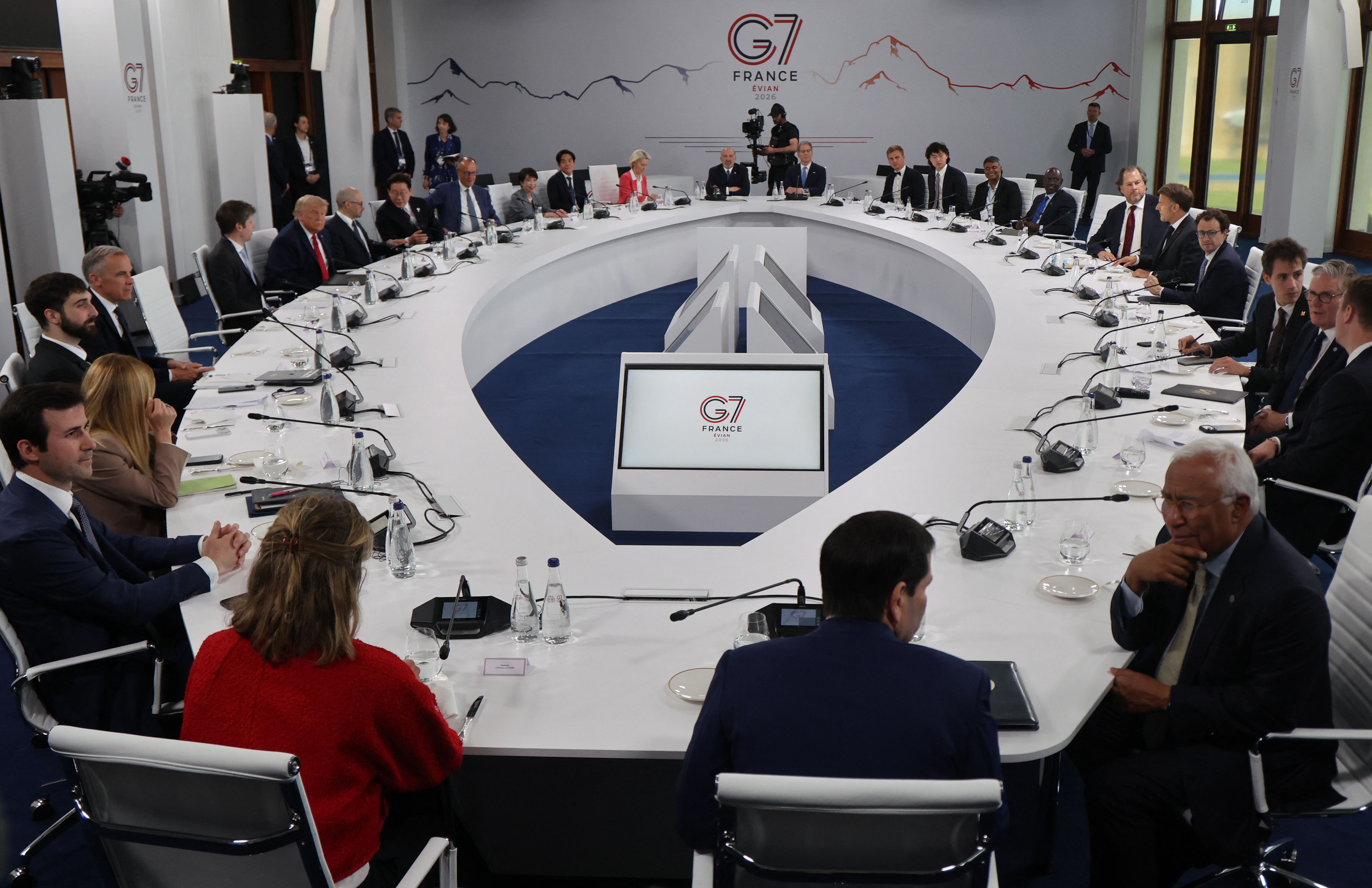 People sit around a round white conference table with mic and water bottles. A screen showing "G7" is in the center.