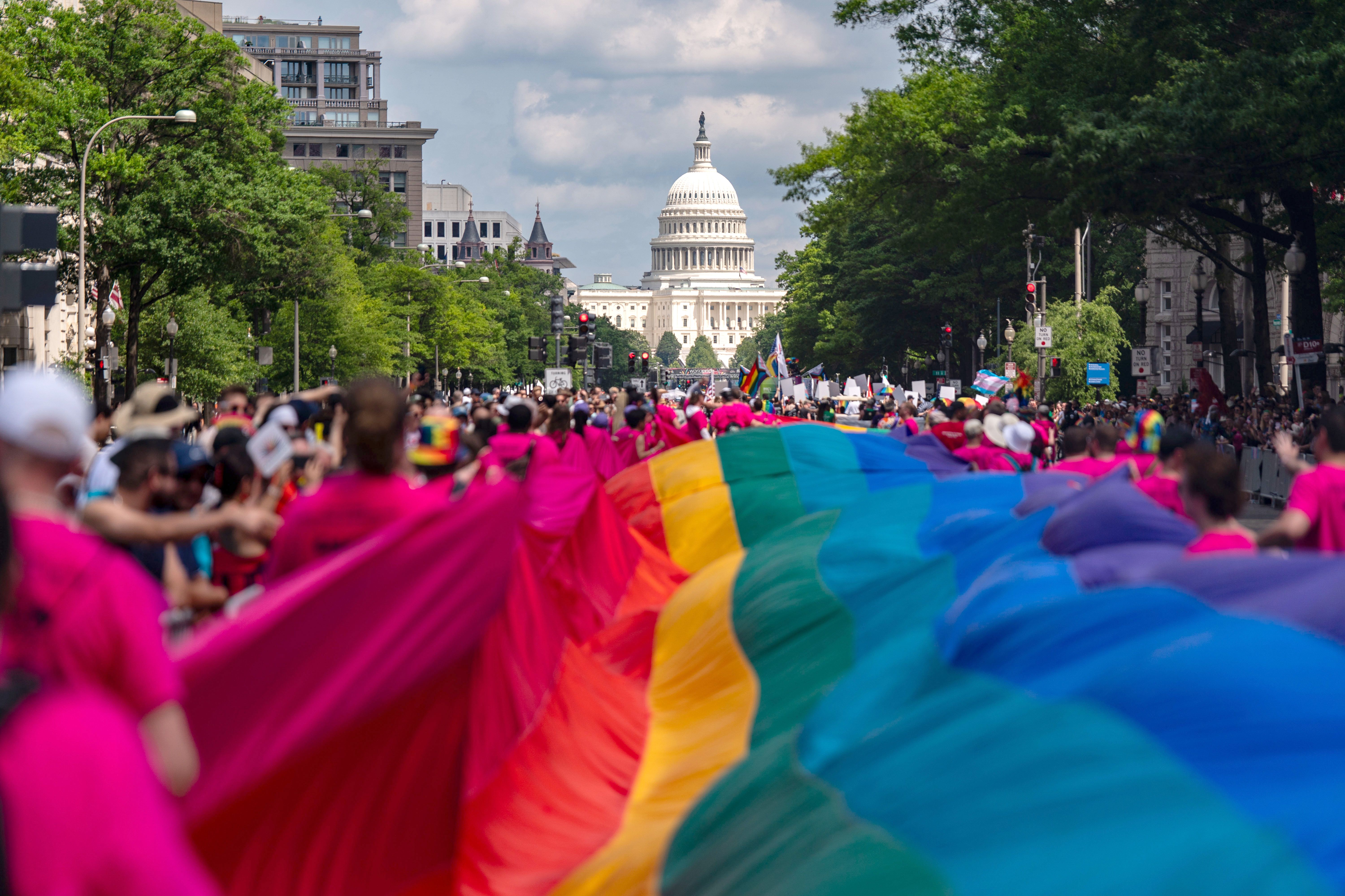 A large crowd in pink shirts carries a giant rainbow pride flag down a tree-lined street toward the U.S. Capitol building under a partly cloudy sky.