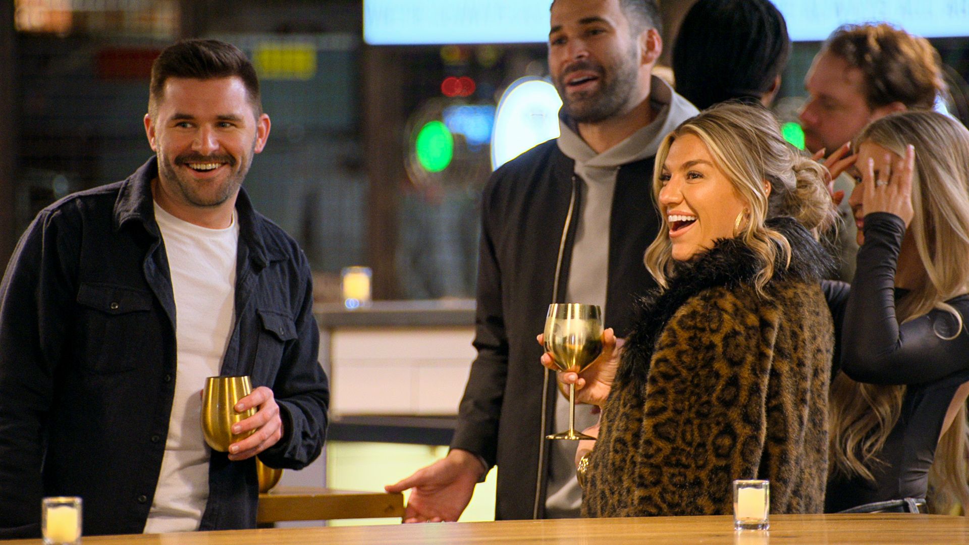 Smiling people at a bar holding golden cups, including a man with a mustache in a black jacket and a woman in a leopard fur coat laughing, with blurred background lights.