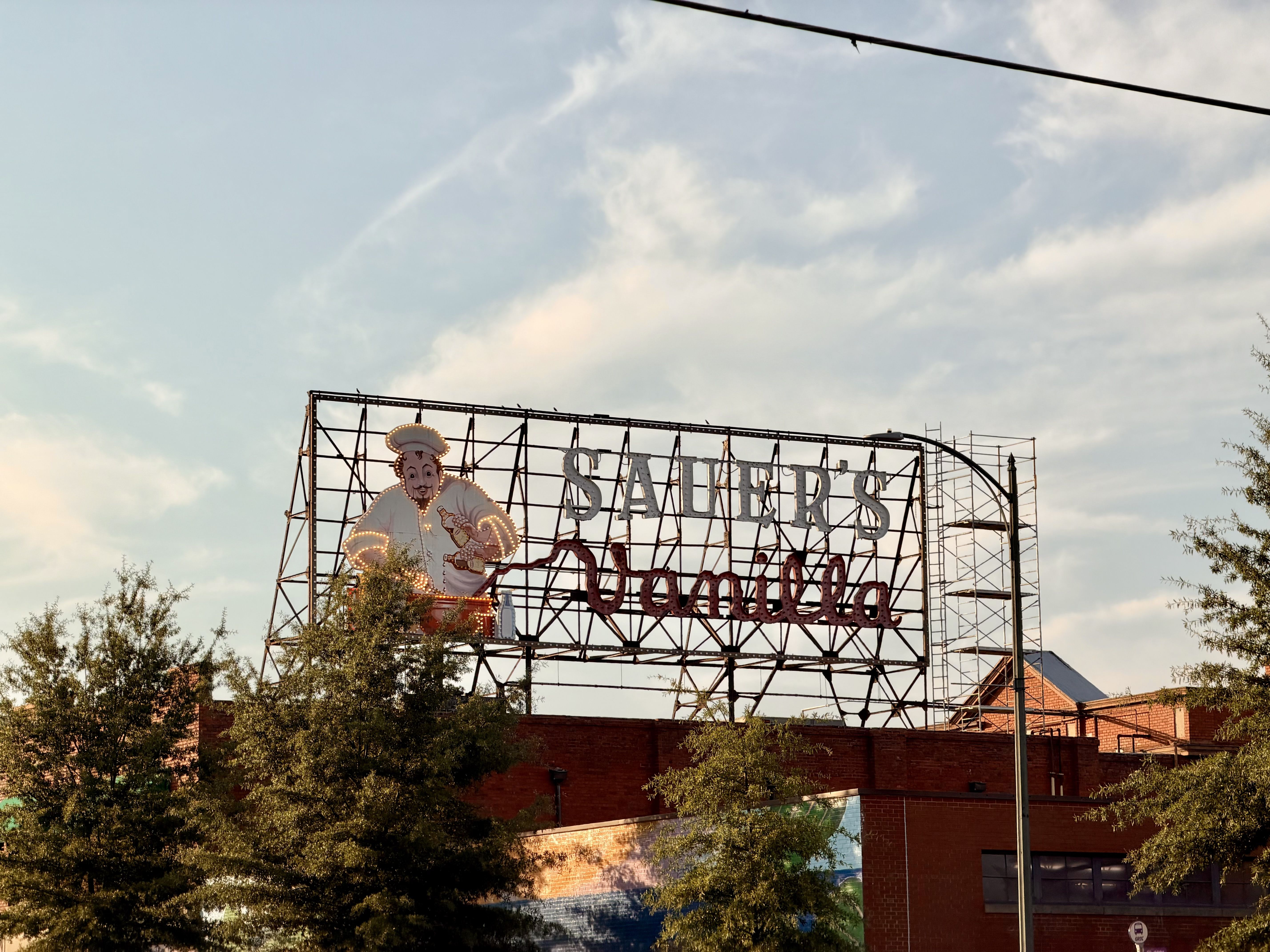 Large open metal sign frame with illuminated letters reading "SAUER'S" and a cartoon chef, mounted above a brick building; trees in the foreground and a blue sky.
