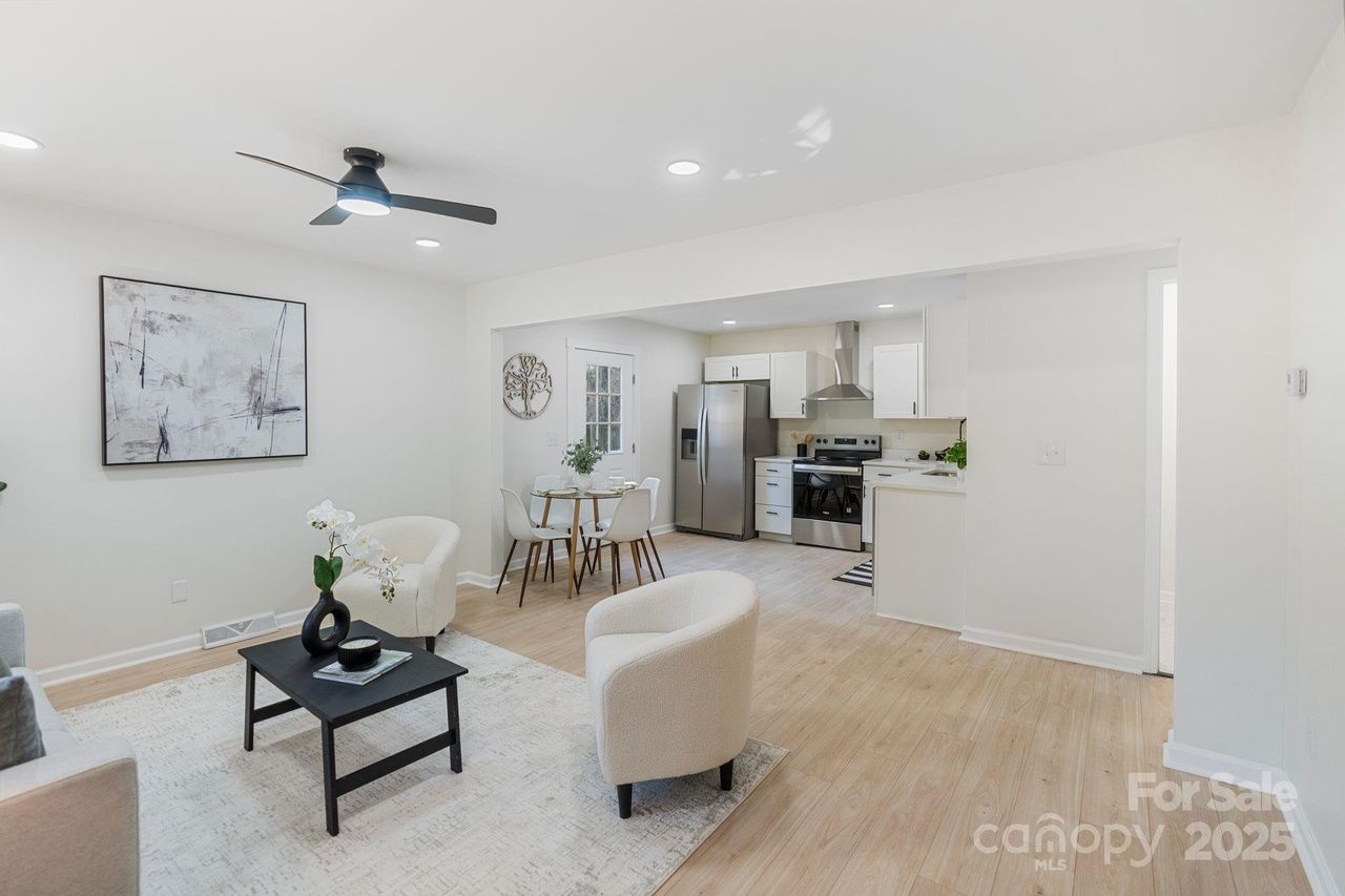 Bright open living room and kitchen with light wood flooring, white walls, white armchairs, black coffee table, abstract wall art, and stainless steel appliances. Ceiling fan overhead.