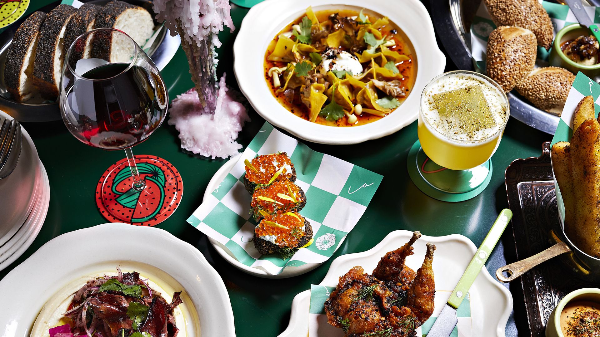 A table with a variety of dishes including sliced bread, a glass of red wine, a yellow cocktail, a bowl of soup with greens, bruschetta topped with red caviar, roasted quail, and seasoned fried sticks.