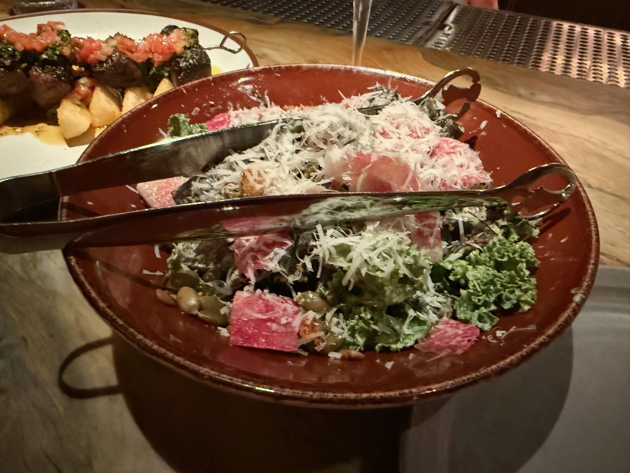 Brown bowl with salad containing kale, lentils, cubes of pink meat, and shredded white cheese, with silver tongs placed on top; blurry skewer with diced meat in background.