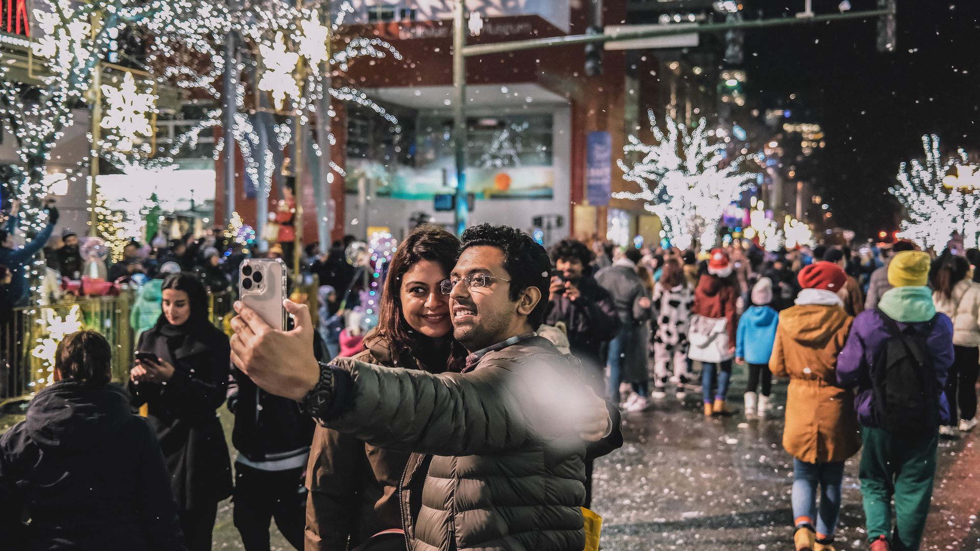 A man and a woman smile for a selfie on a street decorated for the winter holidays. 