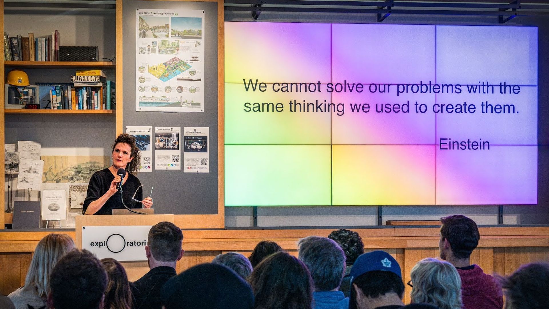 Speaker at a wooden podium in a classroom; a large colorful grid screen displays Einstein's quote: "We cannot solve our problems with the same thinking we used to create them." Audience watches.