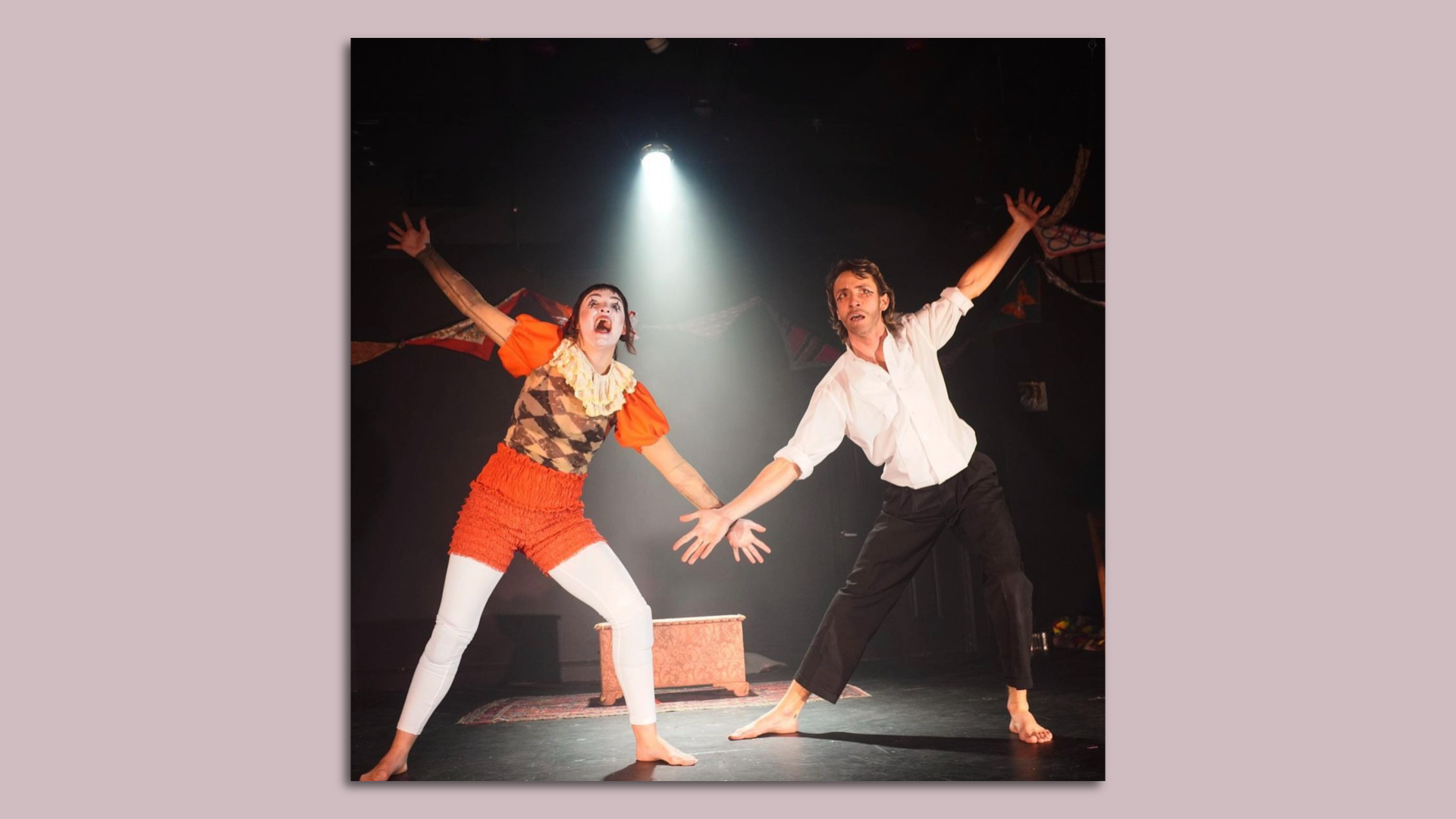 Photo of two clown performers holding their arms out on a stage. The one on the left is in orange clown make-up and clothes.