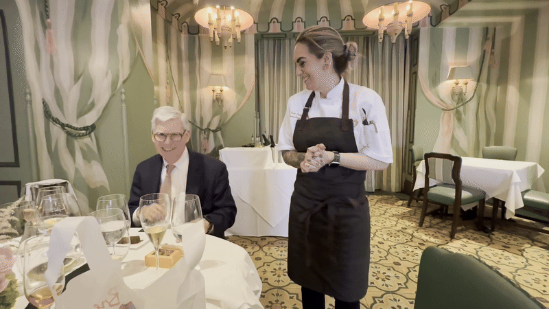 A woman in a chef's coat and apron smiles next to a man who's seated and smiling.