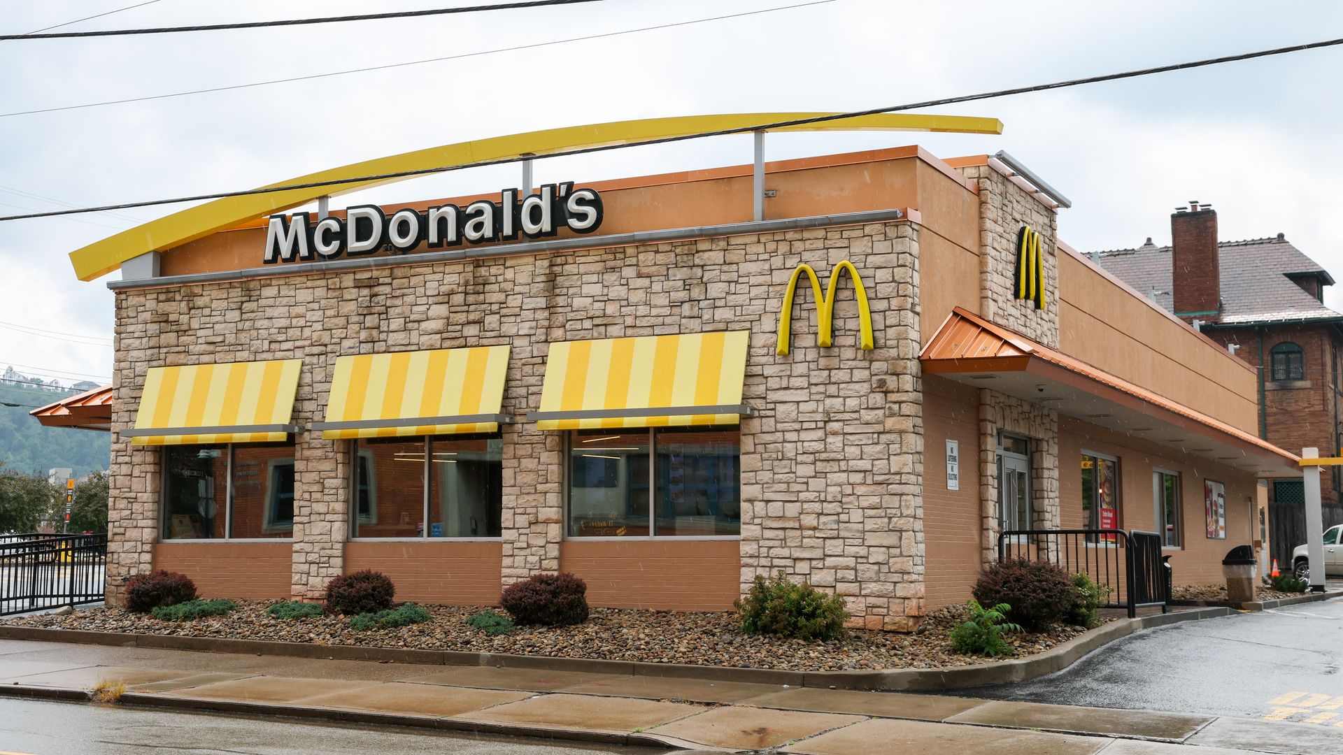 McDonald's fast-food restaurant with a stone facade, a large McDonald's sign, and the yellow arches logo on the wall. Yellow striped awnings, damp pavement, and a cloudy sky.