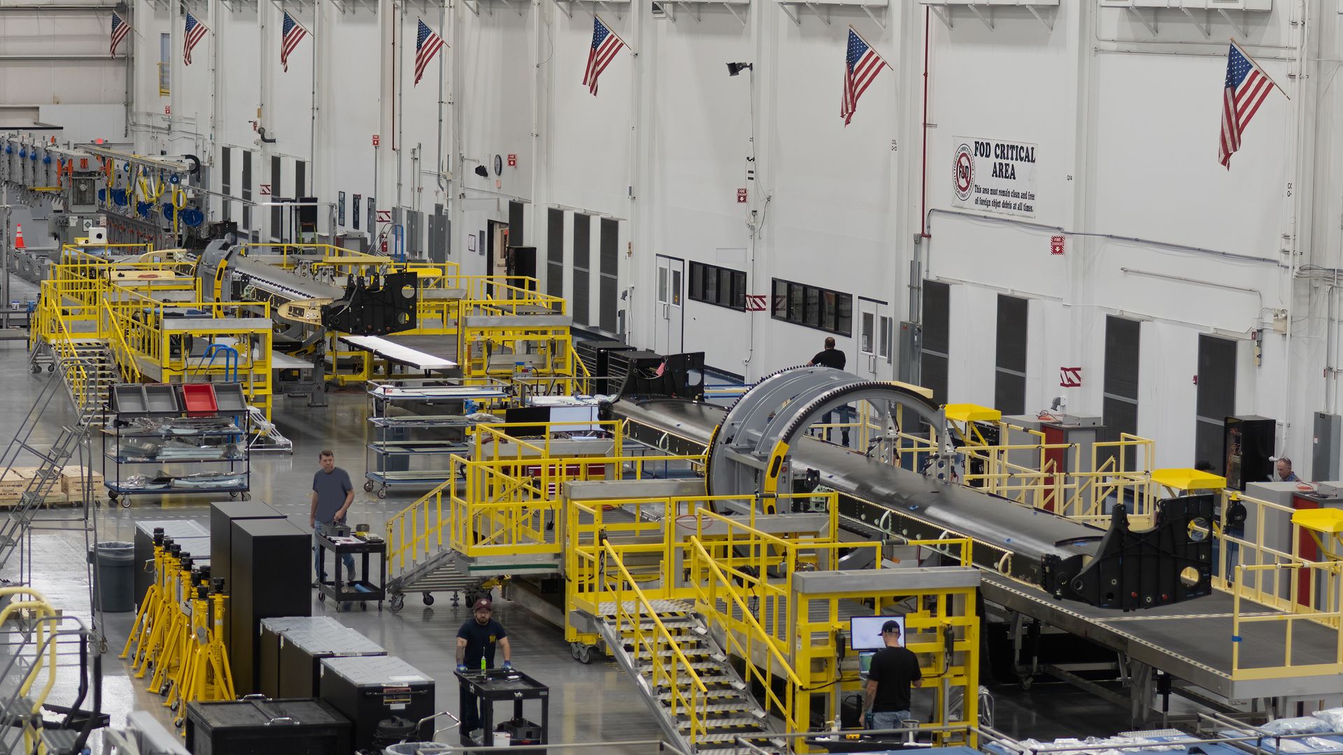 Inside a large aerospace factory with white walls, yellow safety rails, and conveyor systems. American flags hang from the white walls; workers walk near machinery and a curved aircraft parts.