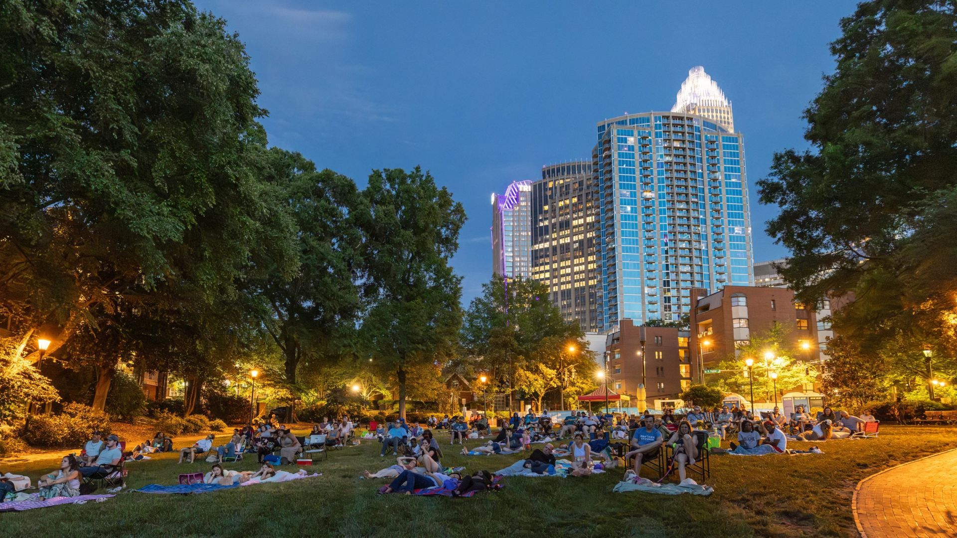 Dusk city park filled with people lounging on blankets, enjoying an outdoor gathering under leafy trees. In the background, modern blue glass skyscrapers glow, with a lit, crown-topped tower visible.
