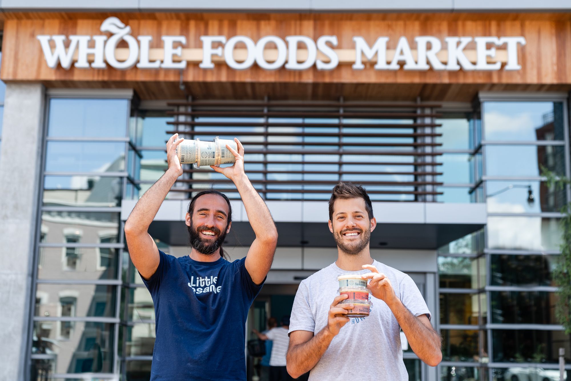 Two smiling men standing outside a Whole Foods Market holding containers of Little Sesame hummus, one in a navy blue shirt and the other in a light gray shirt.