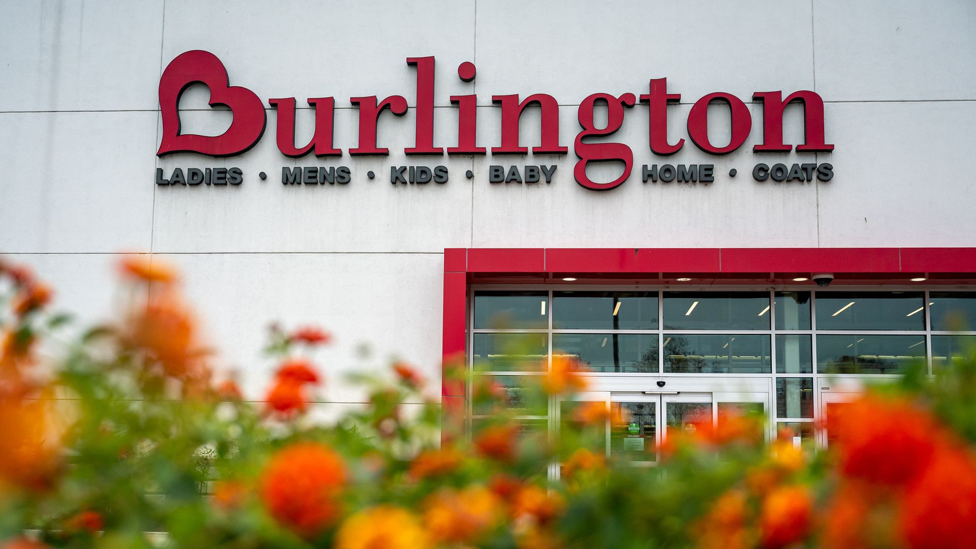 Sign reading "Burlington" in large red letters on a pale wall, with gray category words beneath, reading ladies, mens, kids, baby, home, and coats. Foreground orange flowers blur; glass entrance with red framing.