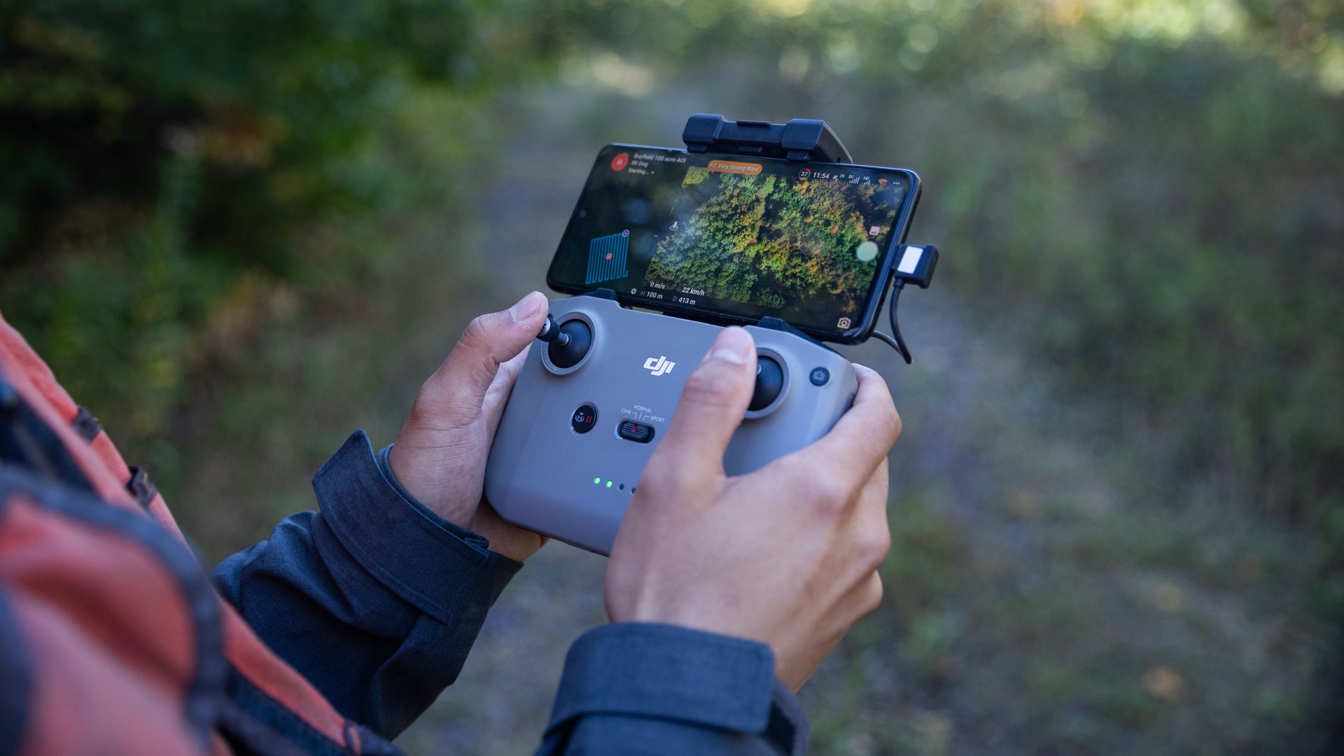 Person holding a gray DJI drone controller with a smartphone showing a forest map and flight details outdoors with blurred greenery in the background.