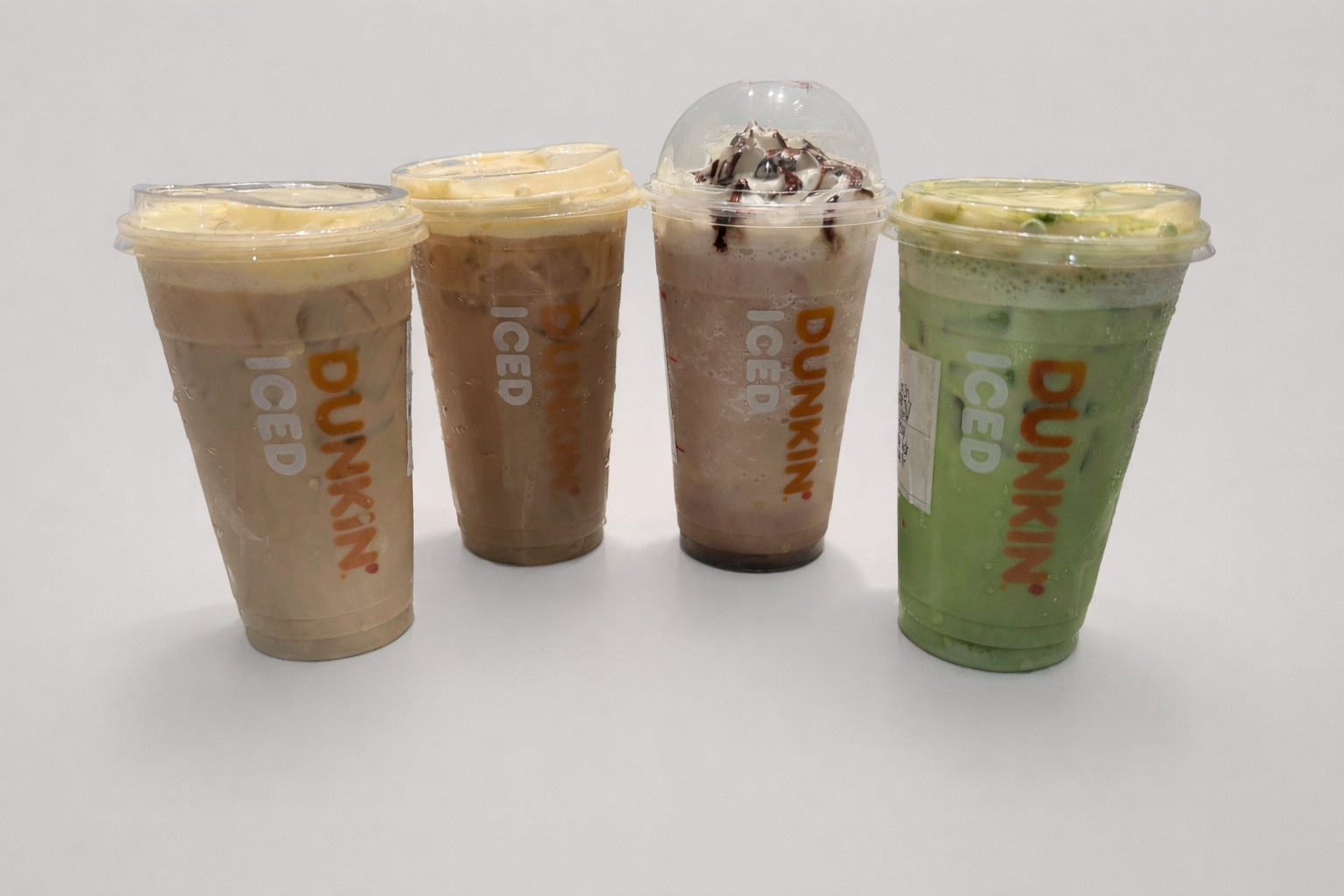 Four Dunkin iced drinks in clear plastic cups with lids, including two creamy light brown coffees, a chocolate drink with whipped cream and chocolate drizzle, and a green matcha latte.