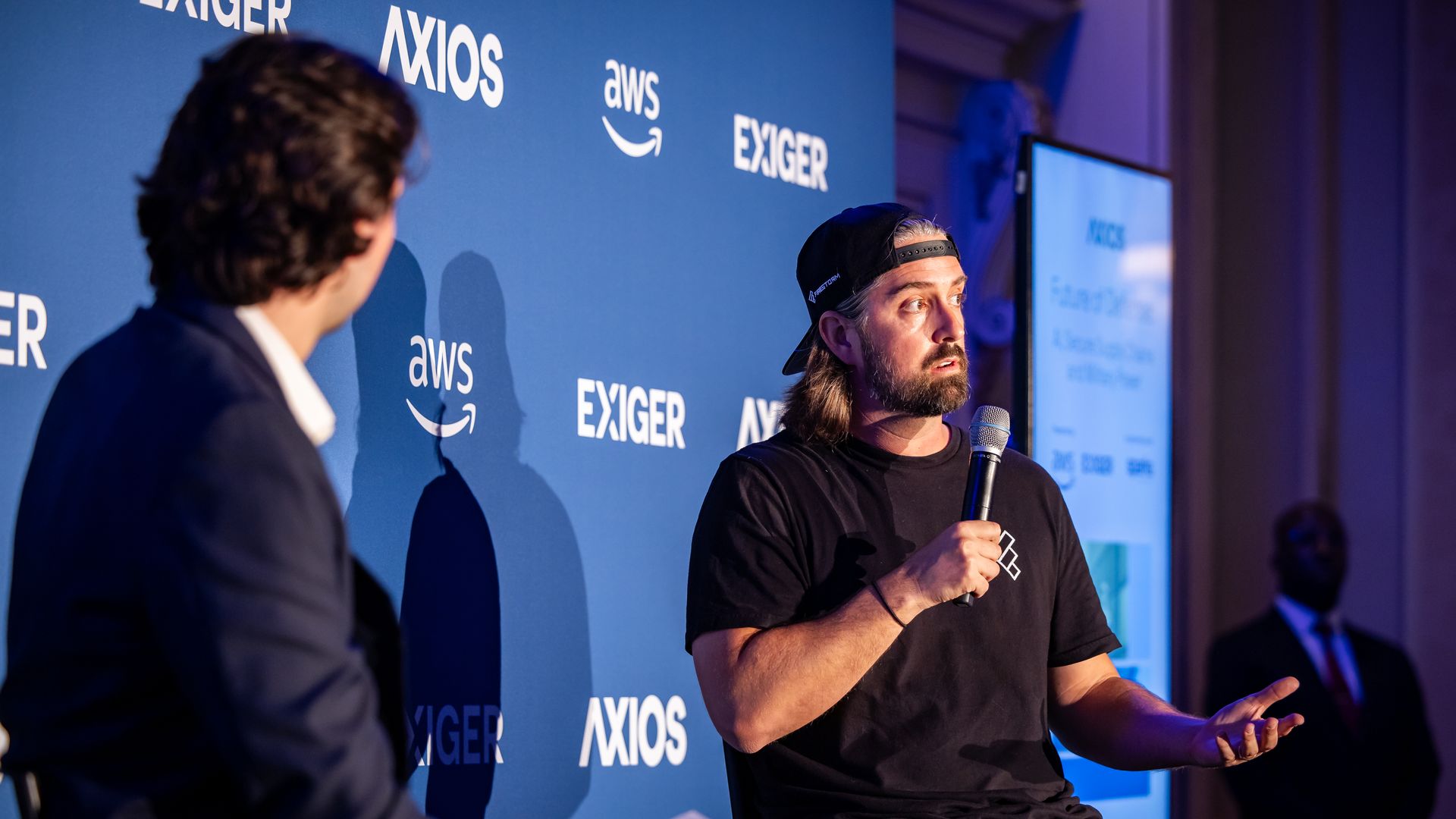 Two men sit on stage with microphones. The backdrop is blue. A man in a suit stands offstage to the right.
