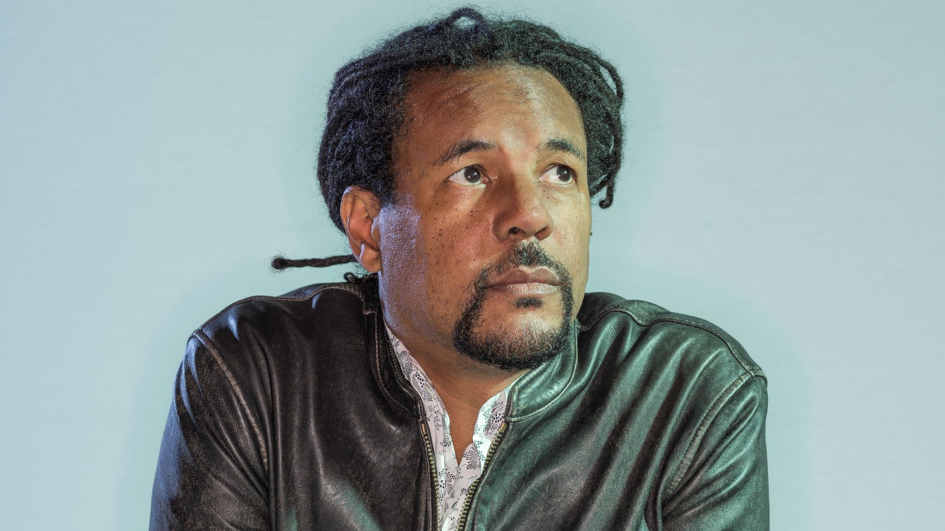 Portrait of a man with short dreadlocks and a goatee, wearing a black leather jacket, leaning on a white-pink pedestal, looking upward against a pale blue backdrop.