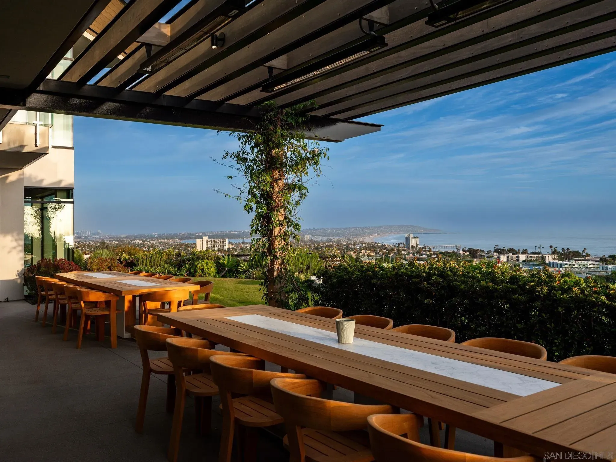 Outdoor terrace with long wooden tables and matching chairs beneath a dark slatted roof. A leafy vine climbs a central tree; city and ocean stretch across the horizon under a clear blue sky.