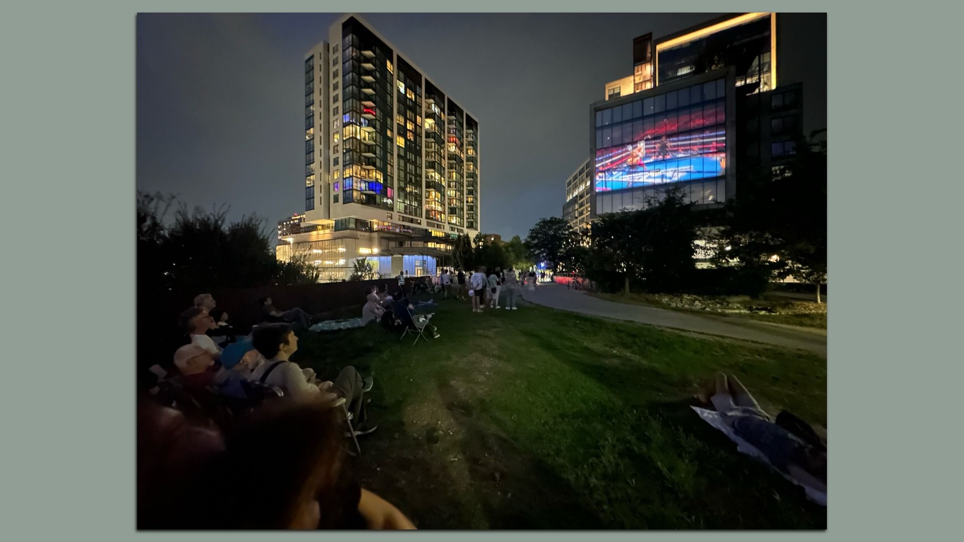 People gathered on grass at night watching a boxing match projected on a large building screen, with tall lit buildings in the background.