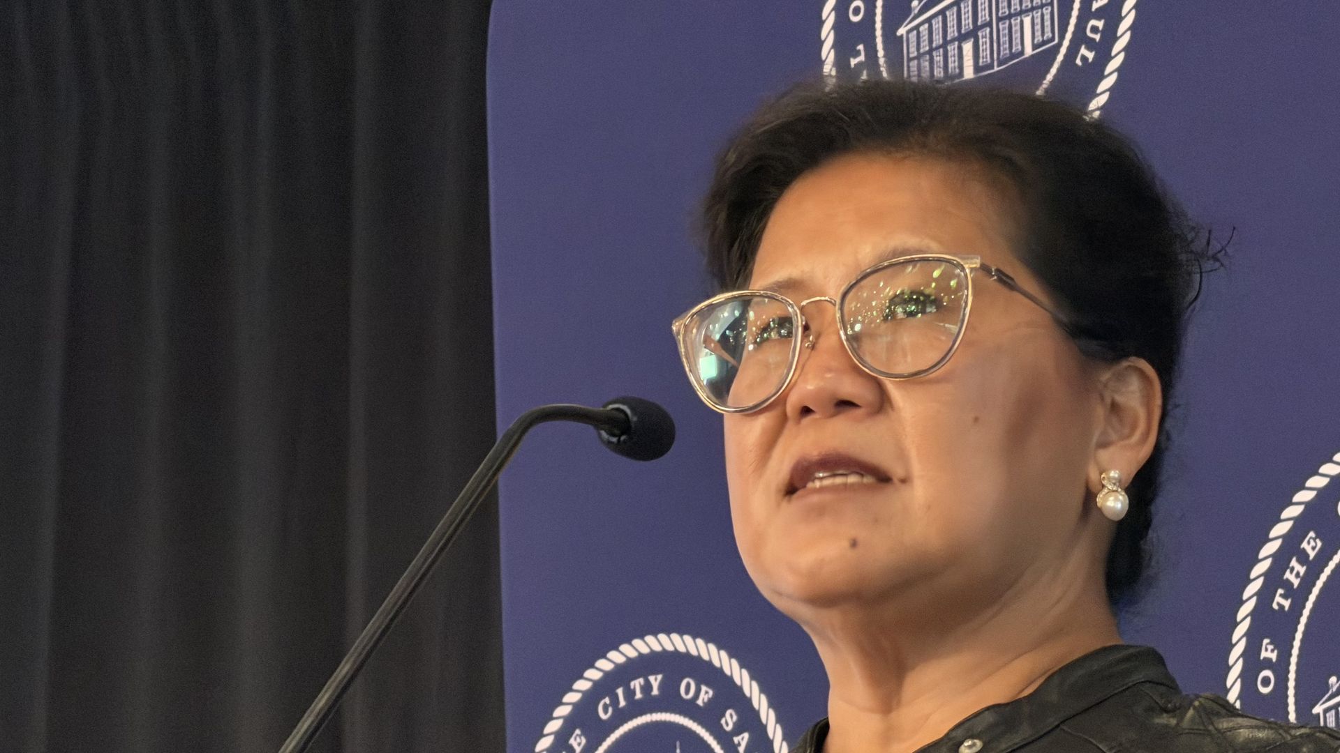 Close-up of a woman with short dark hair and large round glasses speaking into a microphone, against a blue backdrop with a circular seal and rope border.