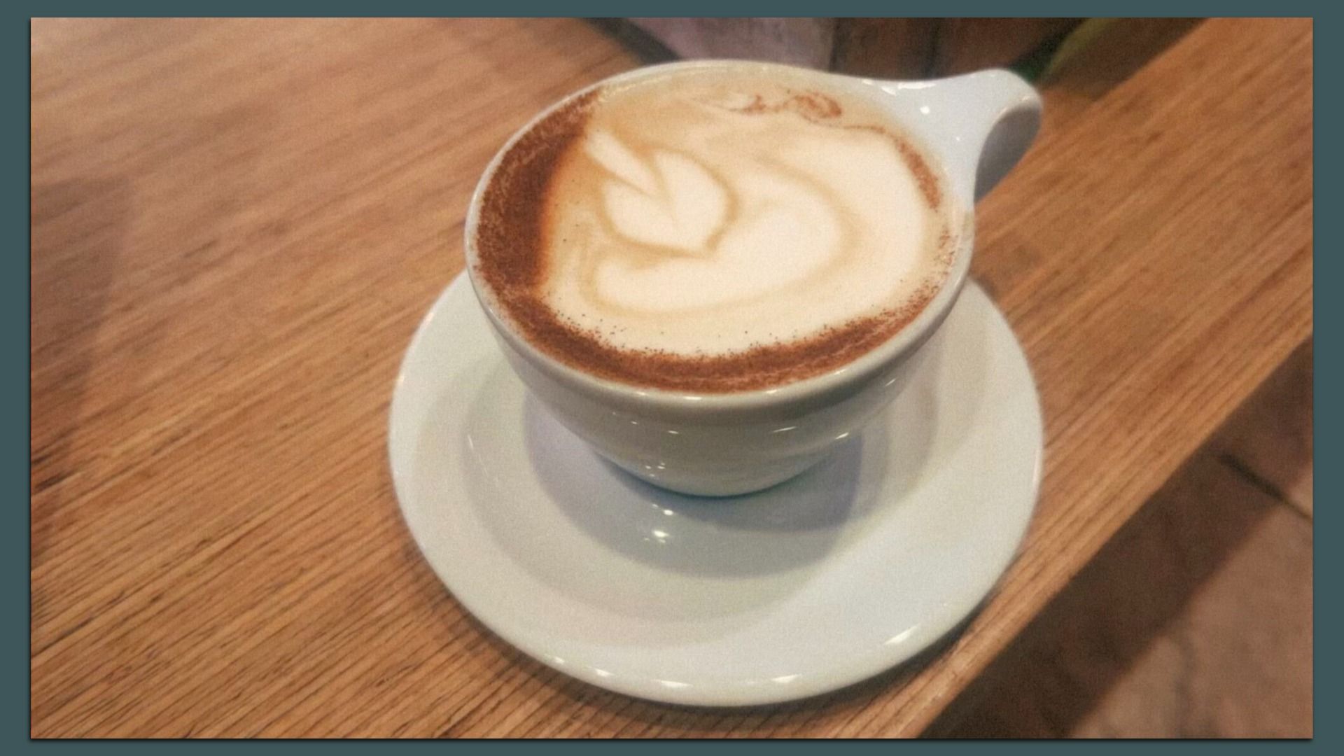 White ceramic cup of cappuccino with heart-shaped latte art and a cocoa-dusted rim, sits on a white saucer on a wooden table.
