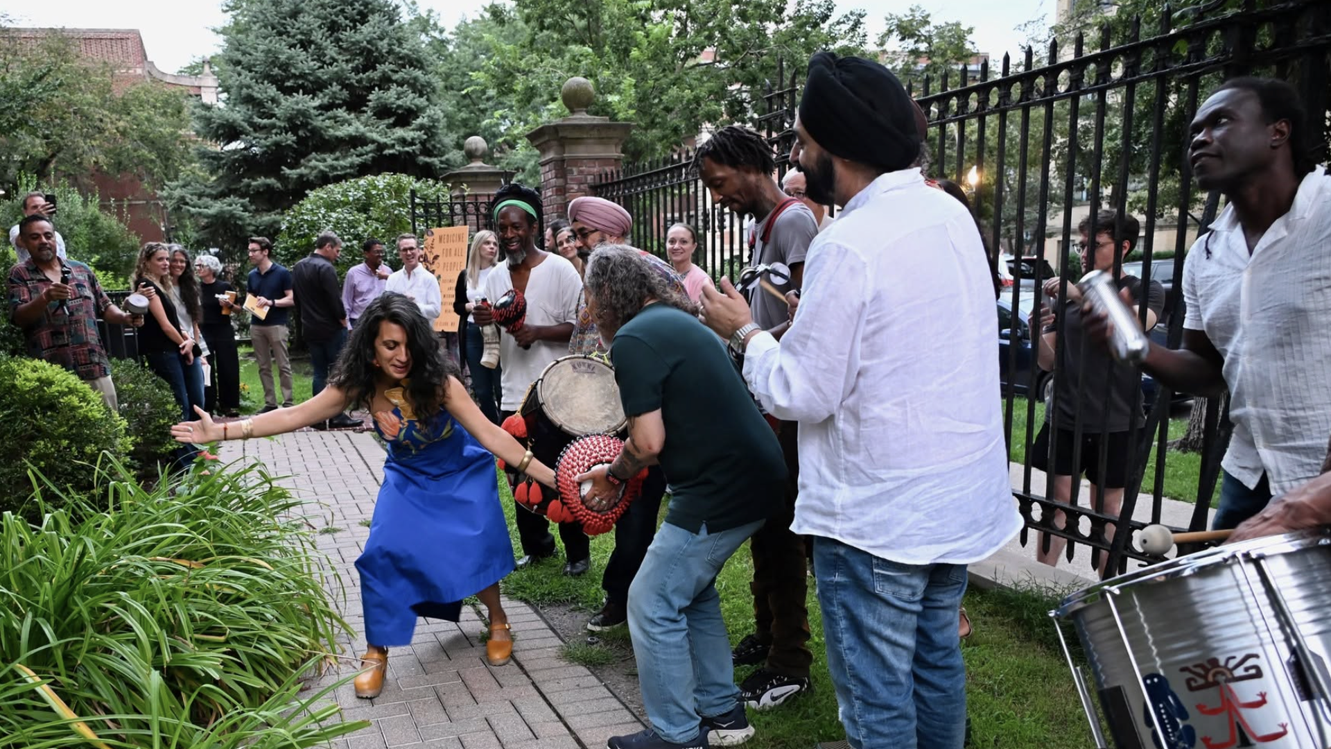 An outdoor street performance with a diverse crowd along a brick path beside a black wrought-iron fence; a woman in a blue dress dances at the front while drummers and musicians play.