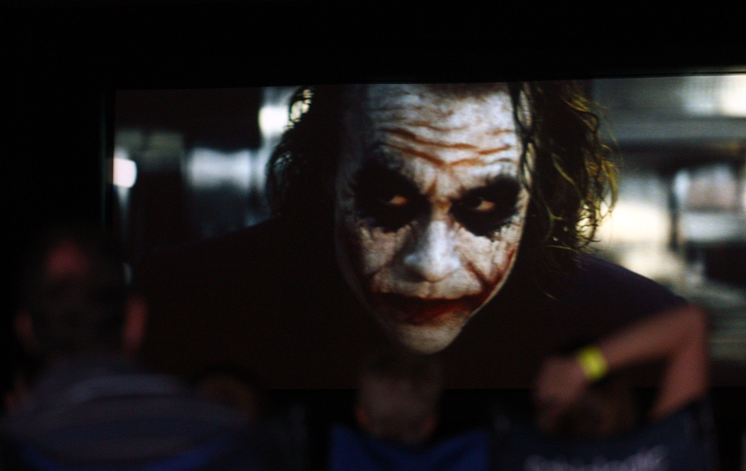 Close-up of a person with white clown makeup, dark eyes, and red scar-like lines across the cheeks, in a dark, blurry background.