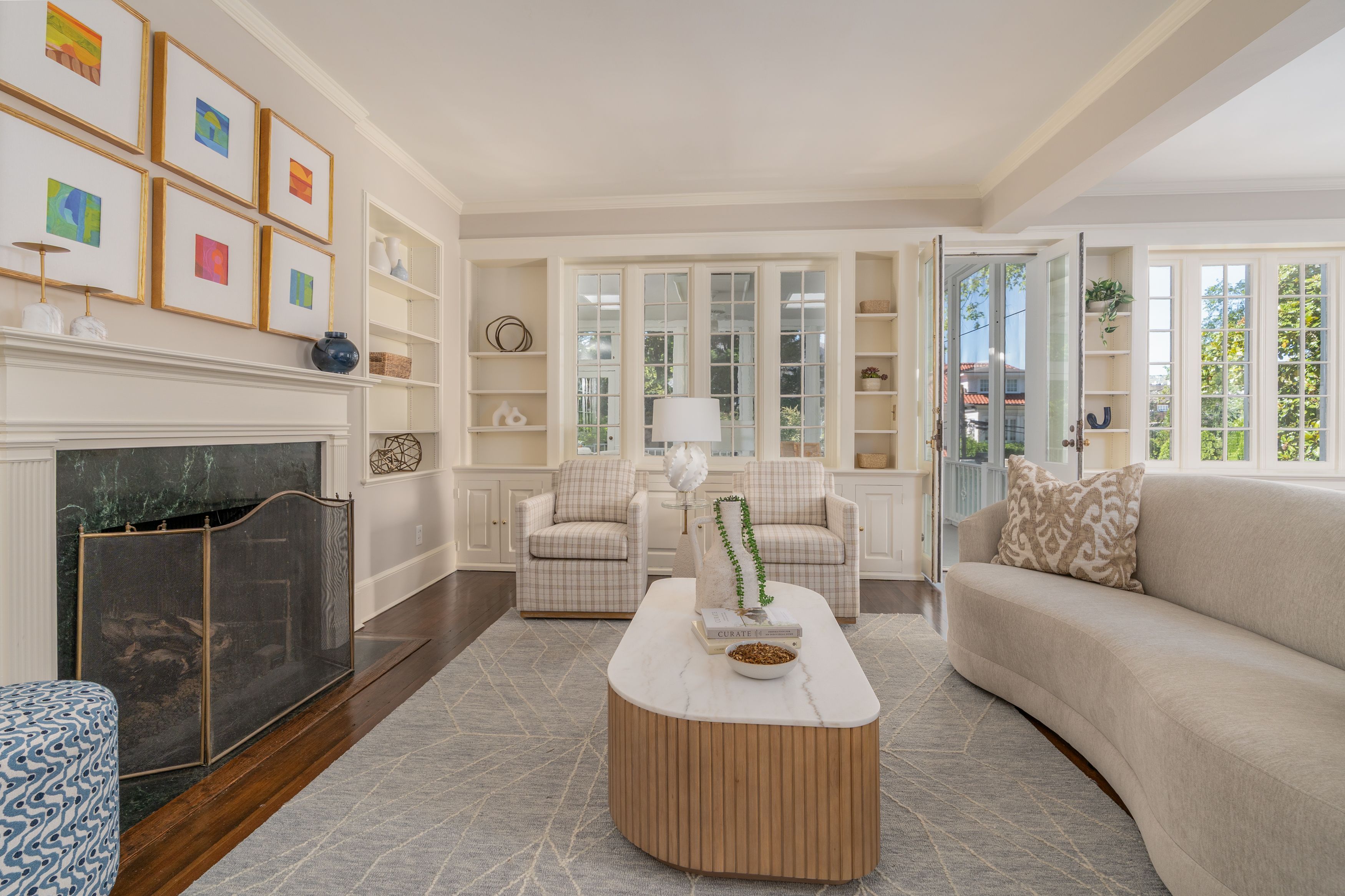Bright living room with white built-in shelves, a fireplace, six gold-framed abstract art pieces above, glass-pane doors, plaid chairs, a beige curved sofa, and a marble-topped coffee table.
