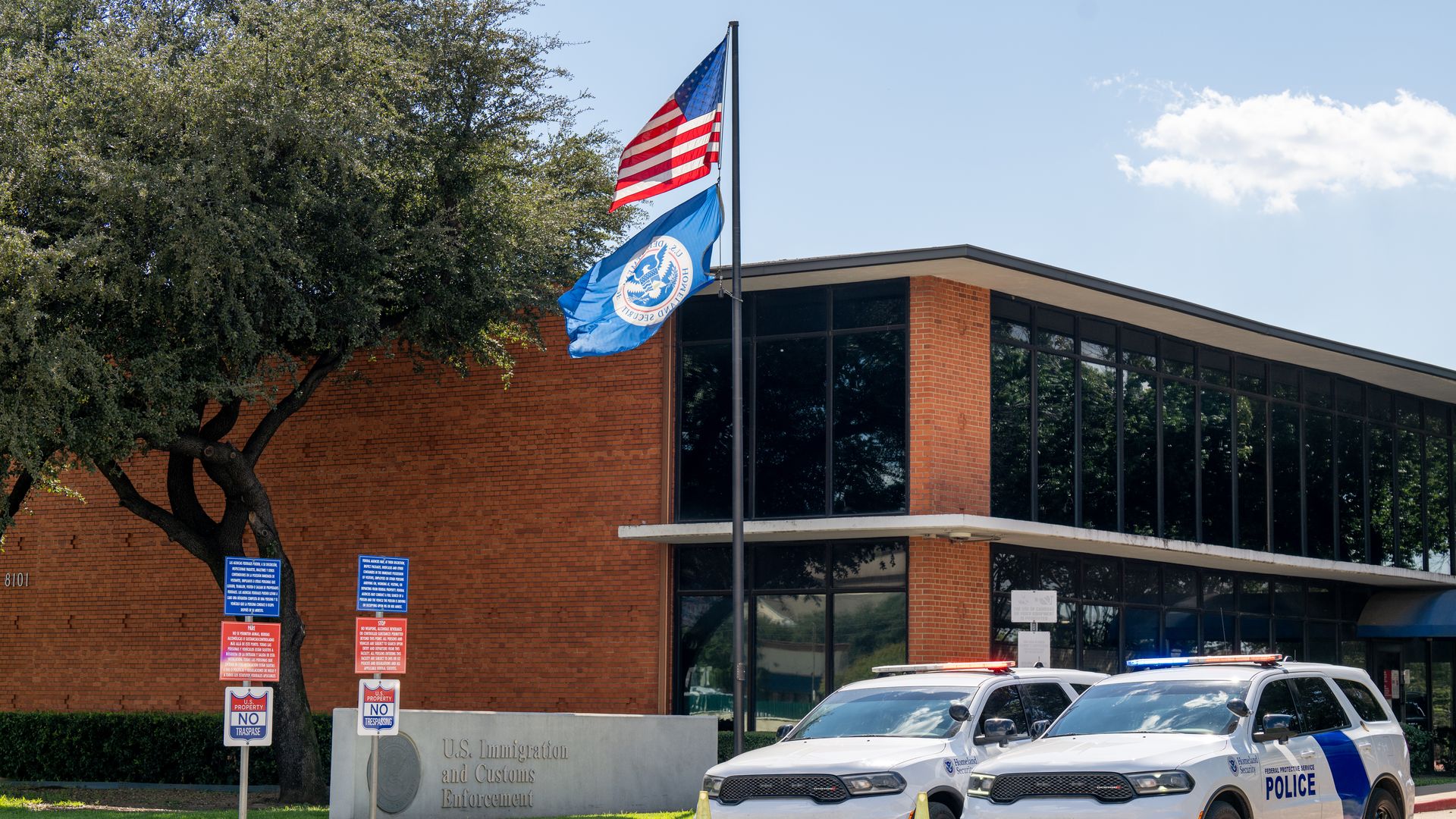 Two white U.S. Immigration and Customs Enforcement police SUVs with flashing lights parked outside a brick government building with the American and DHS flags flying.
