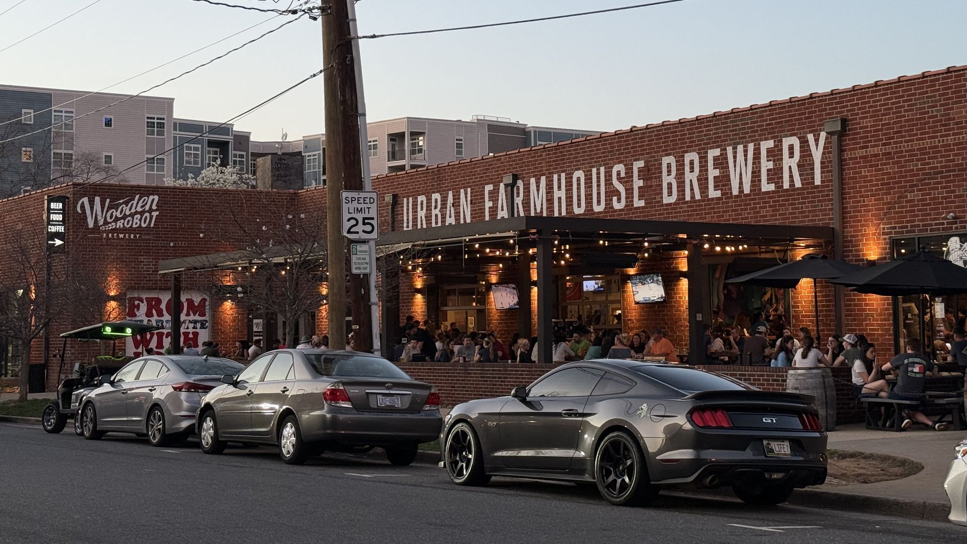 Brick Urban Farmhouse Brewery with a busy outdoor patio and string lights; diners fill the seating area. A "Wooden Robot Brewery" sign sits to the left as cars park along the street.