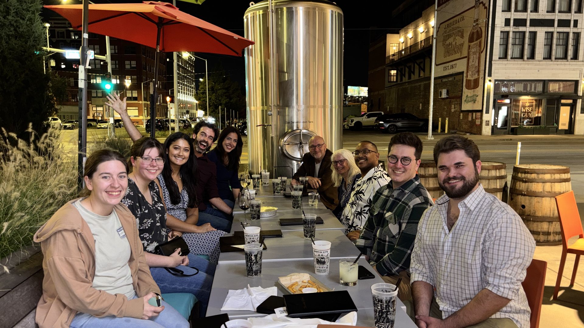 Group of friends seated at a long outdoor table at night under an orange umbrella, smiling with drinks and snacks; city street and a tall stainless tank stand behind them.