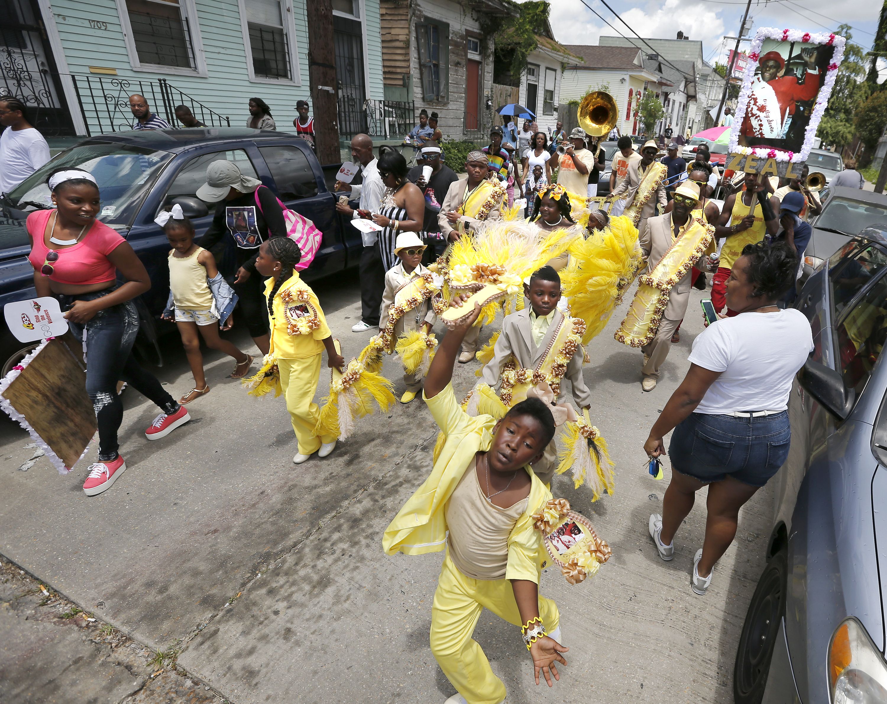 A second-line proceeds down a New Orleans street, where cars are parked on each side. In the roadway are dozens of social aid and pleasure club members who, dressed in yellow and holding fans and other props, dance through the street.
