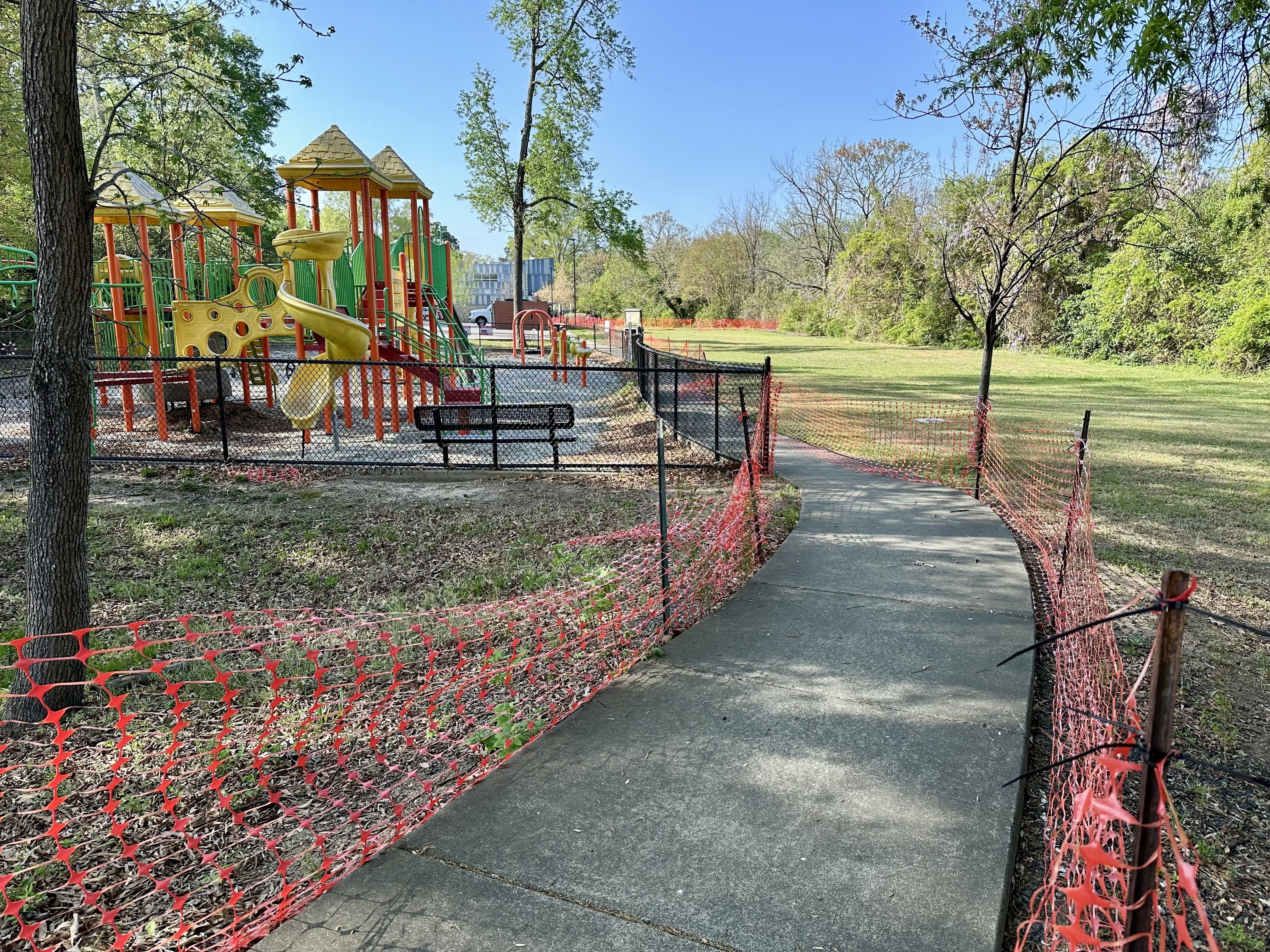 Colorful playground with yellow, green, and red structures behind a black chain-link fence. A curved concrete path lined with orange safety netting winds through a grassy park under a blue sky.