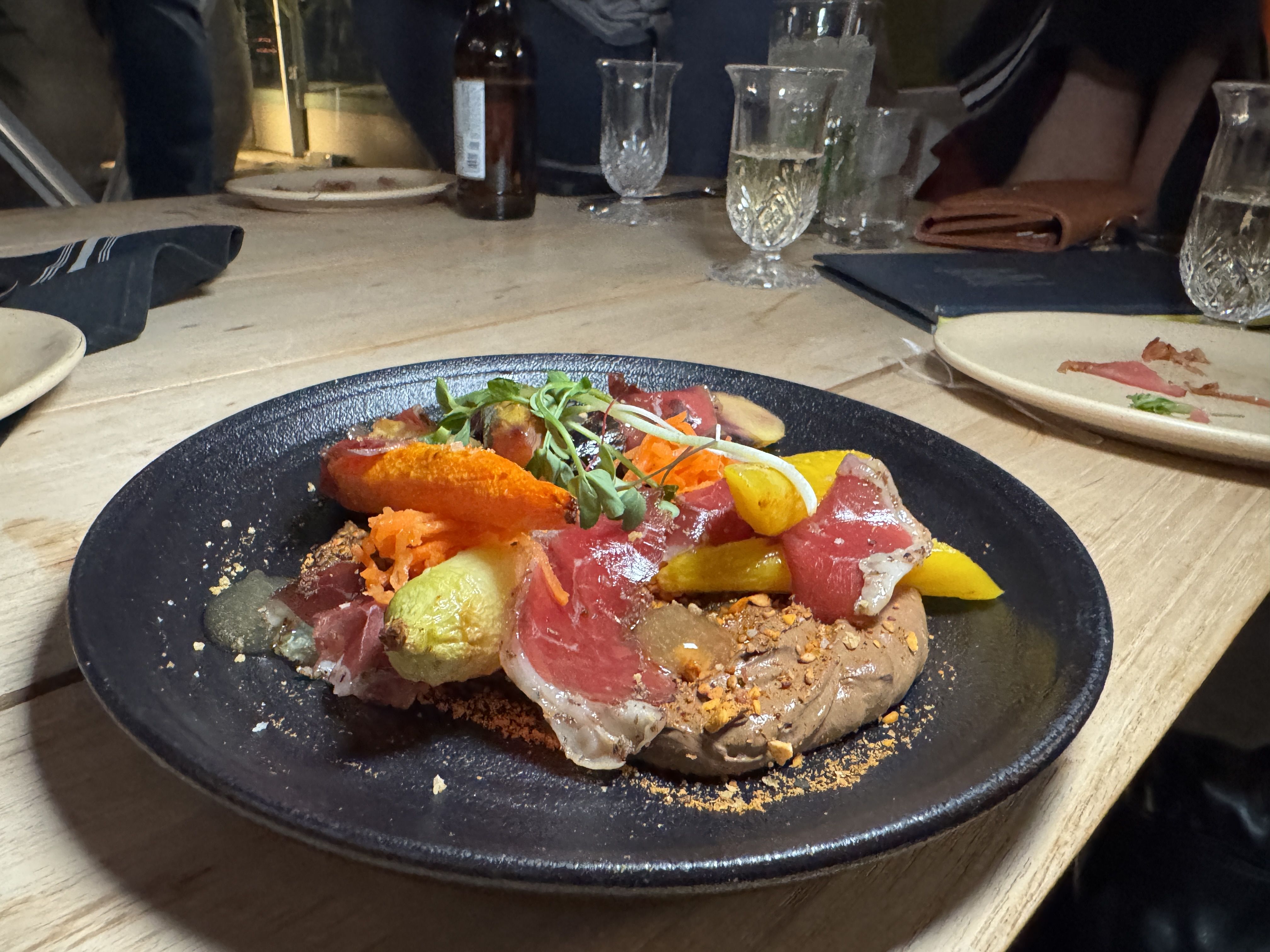 Black plate with colorful gourmet dish including slices of red meat, orange and yellow vegetables, green garnish, and creamy brown sauce on wooden table with drinks and plates in background.