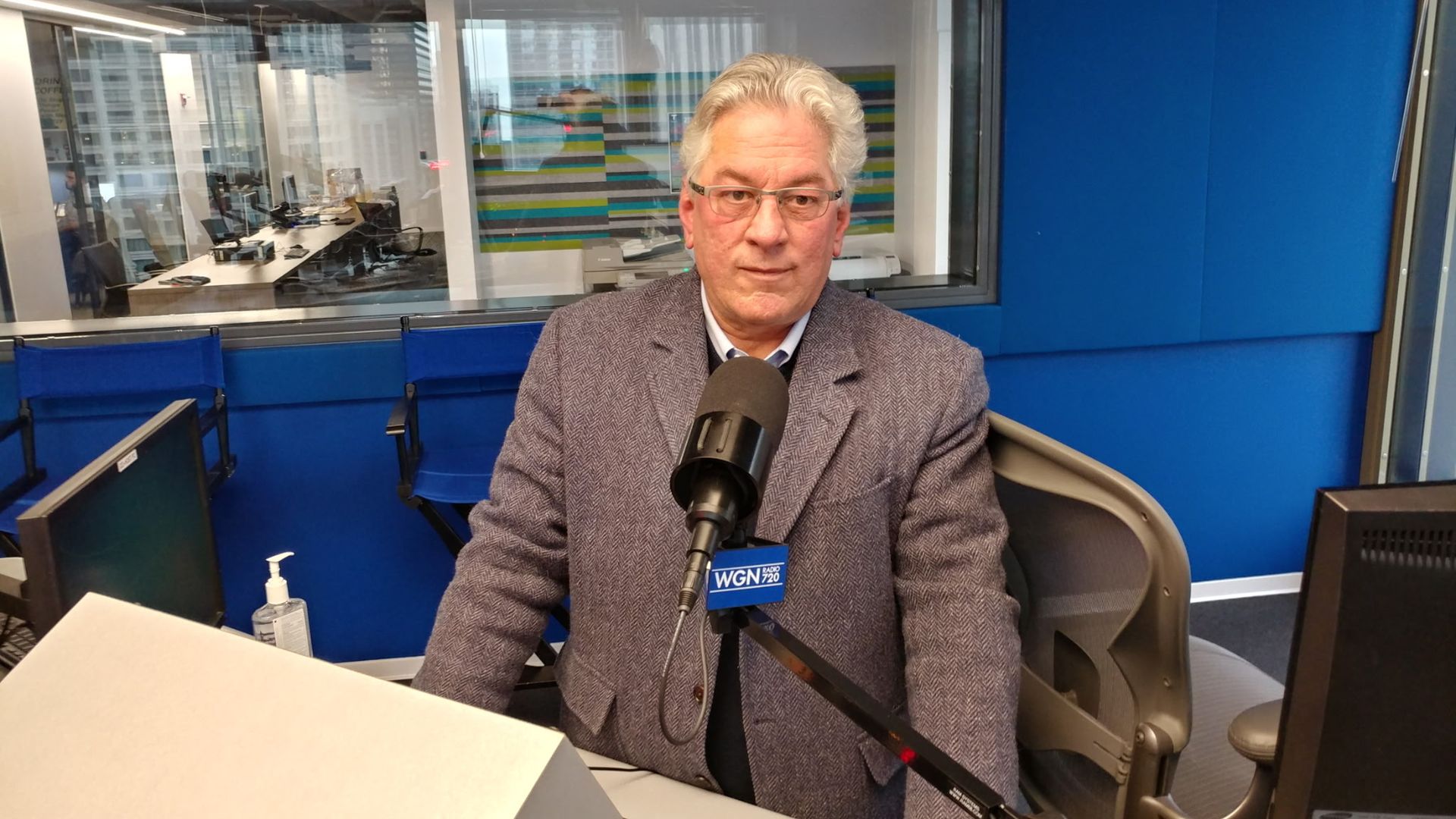 A man in a gray blazer sits in a blue radio studio, speaking into a large microphone labeled WGN 720. A glass window shows a busy control room with computers and striped art.