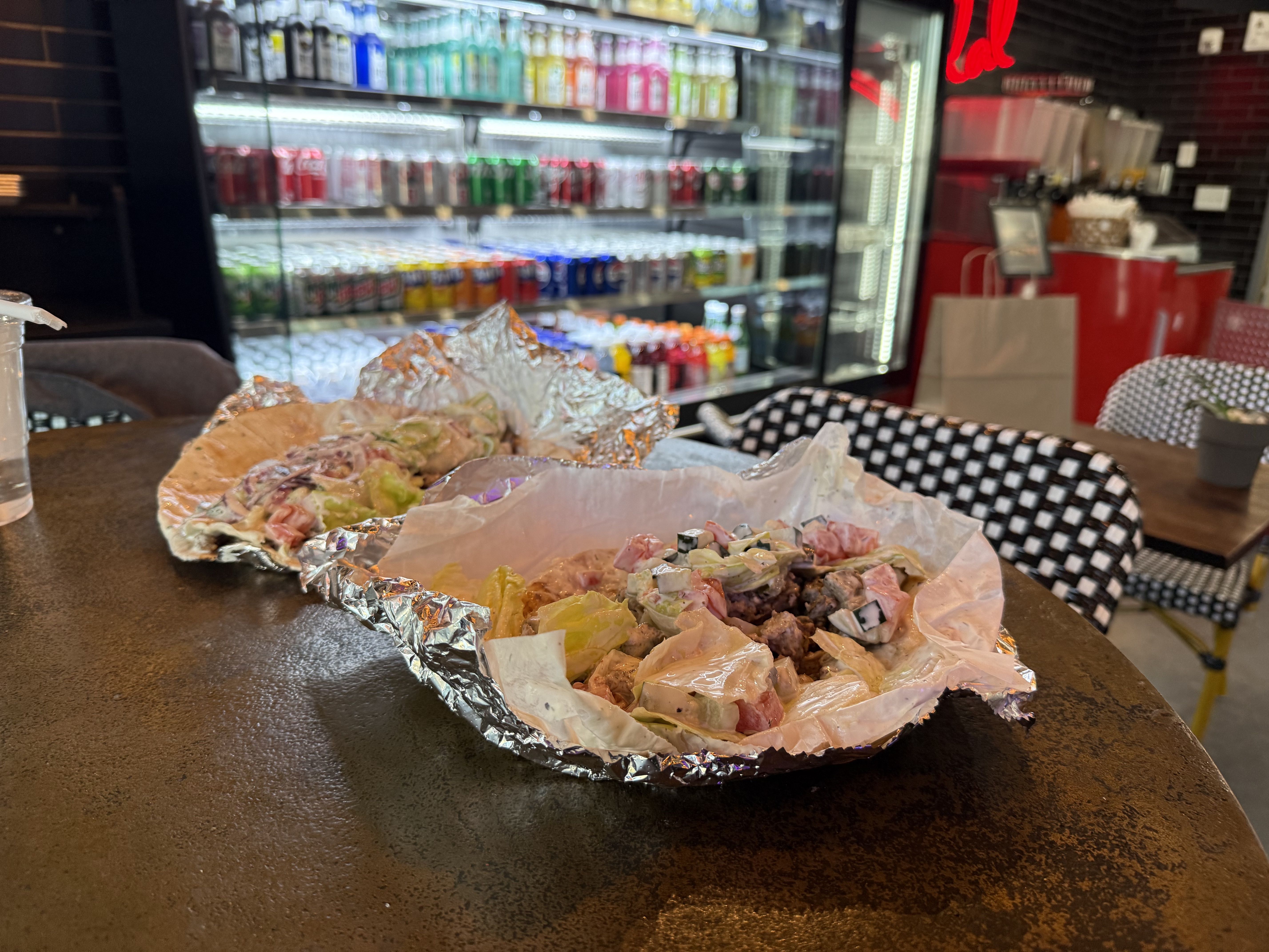 Two partially unwrapped foil sandwiches with lettuce, diced tomatoes, and sauce on a dark table in a casual eatery with a drink fridge and checkered chairs in the background.
