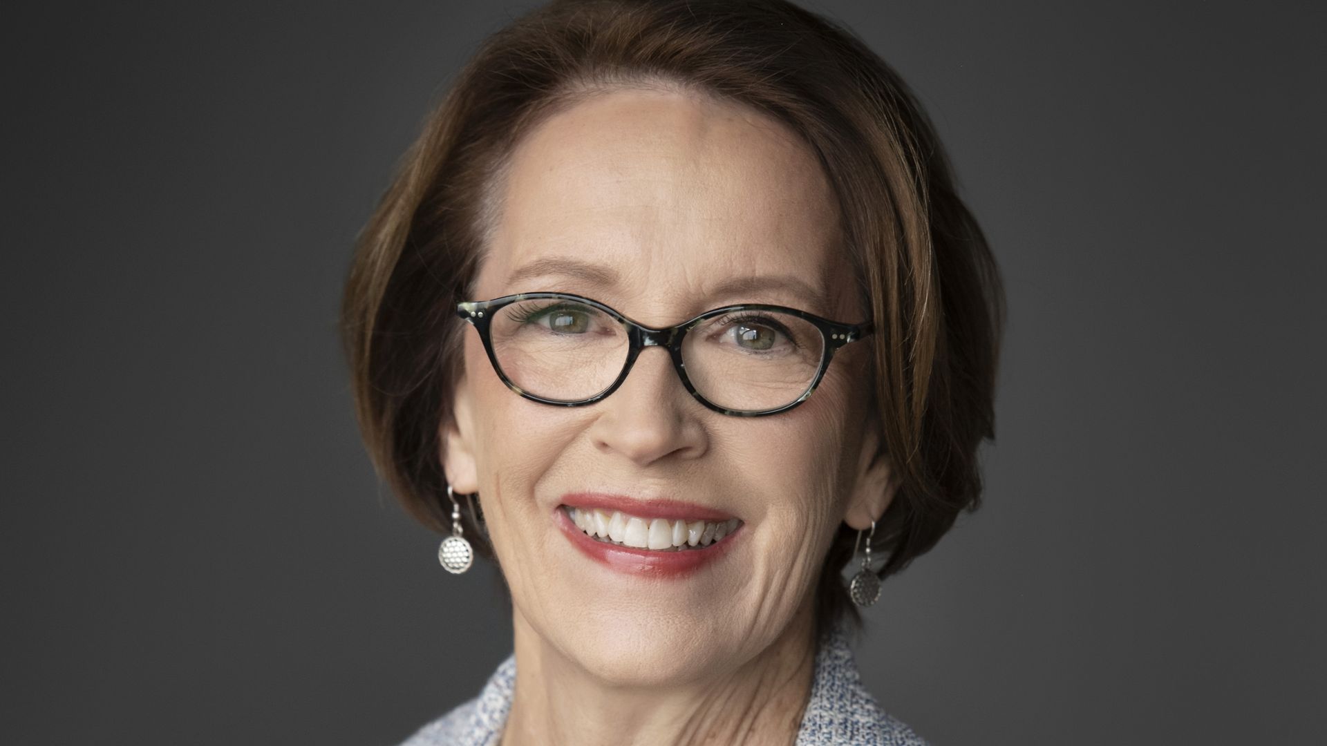 Smiling middle-aged woman with short brown hair and black-framed glasses, wearing a light gray tweed blazer over a black top, against a dark gray background.