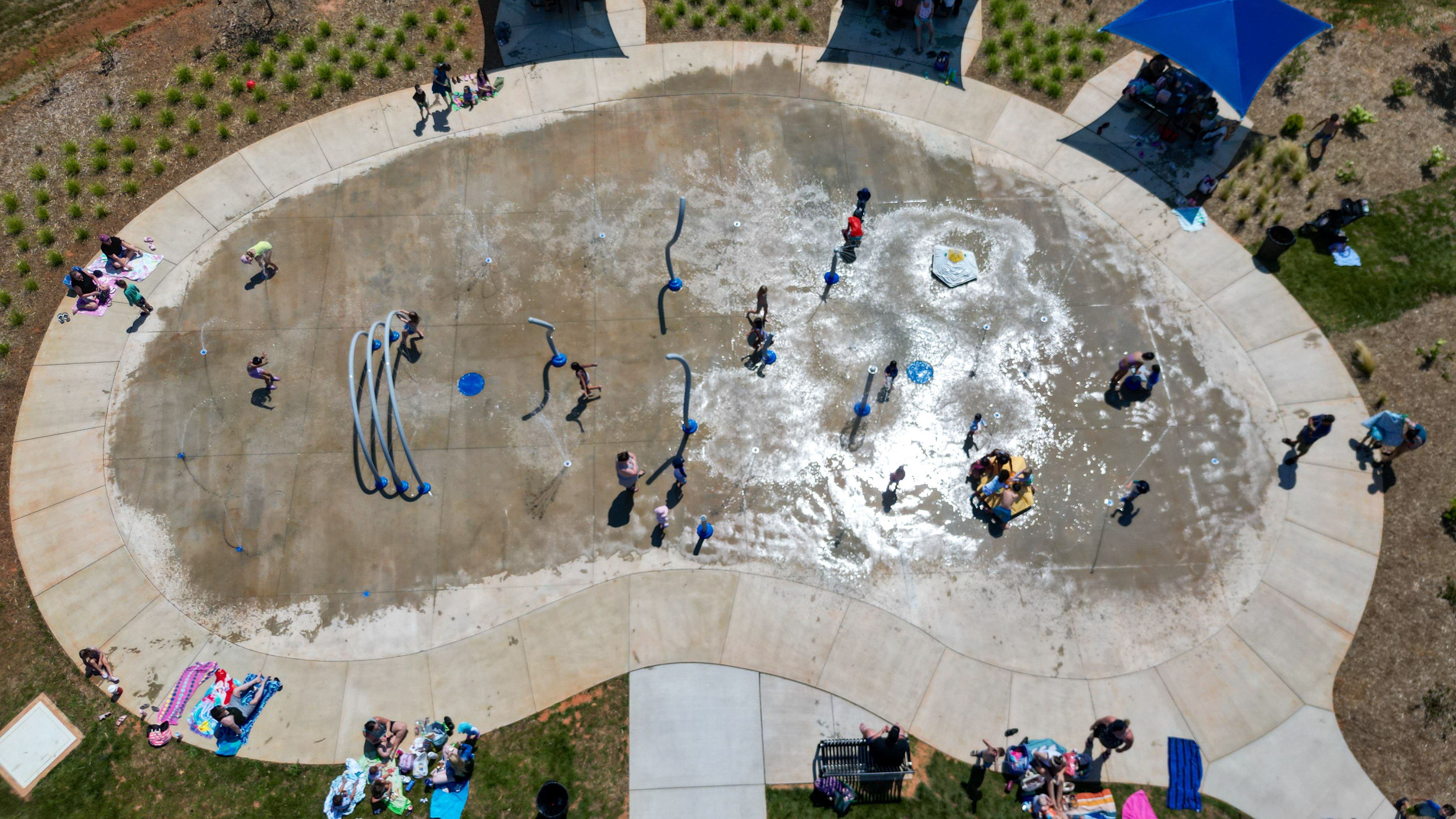 Aerial view of a circular splash pad in a park: kids and adults play in water jets, with towels on the surrounding concrete, and a blue shade canopy on the right.