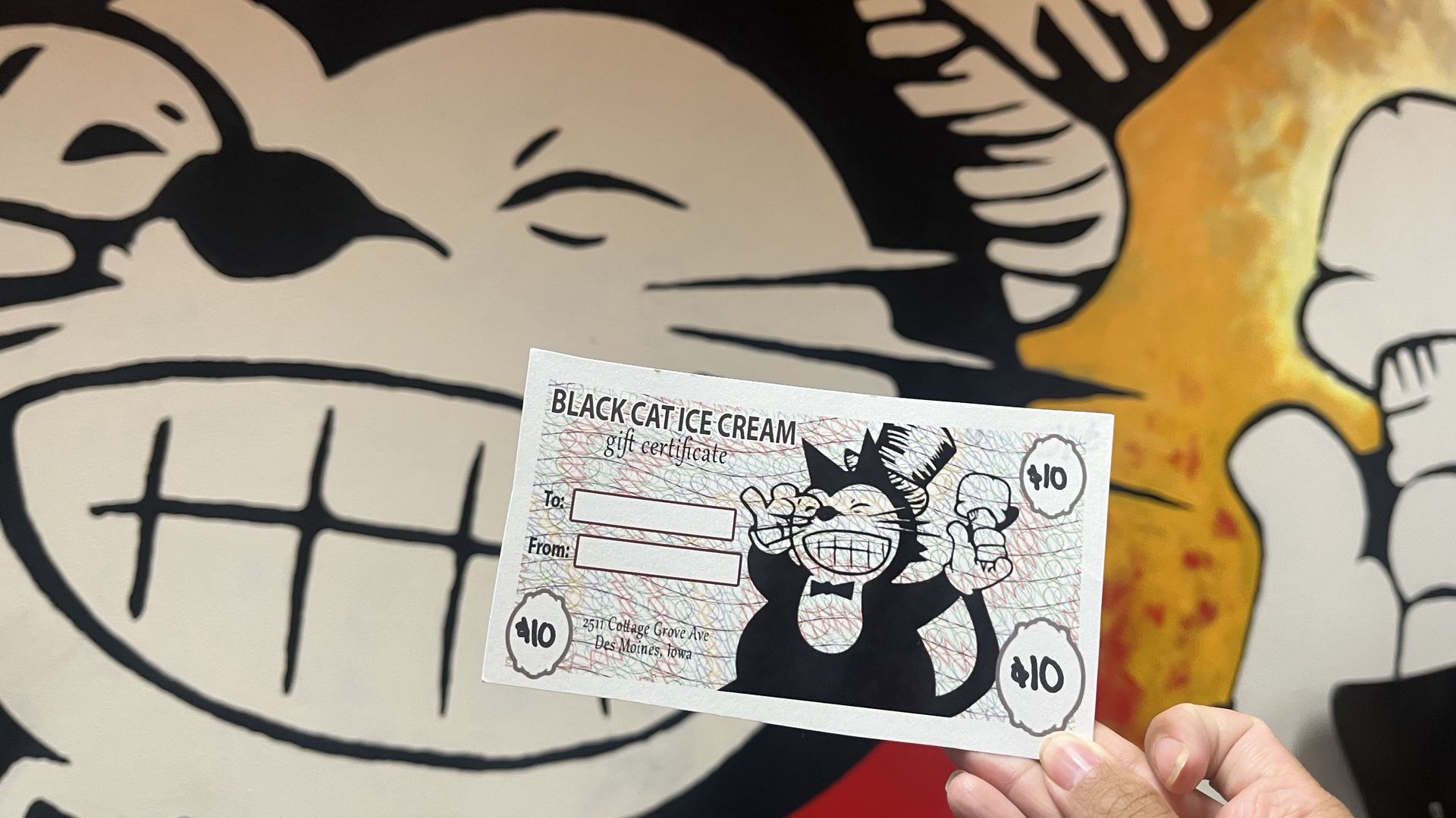 Hand holding a "BLACK CAT ICE CREAM" gift certificate in front of a large beige mural with bold black cartoon lines; a yellow‑orange splash on the right; certificate shows a cat illustration and $10 stamps.