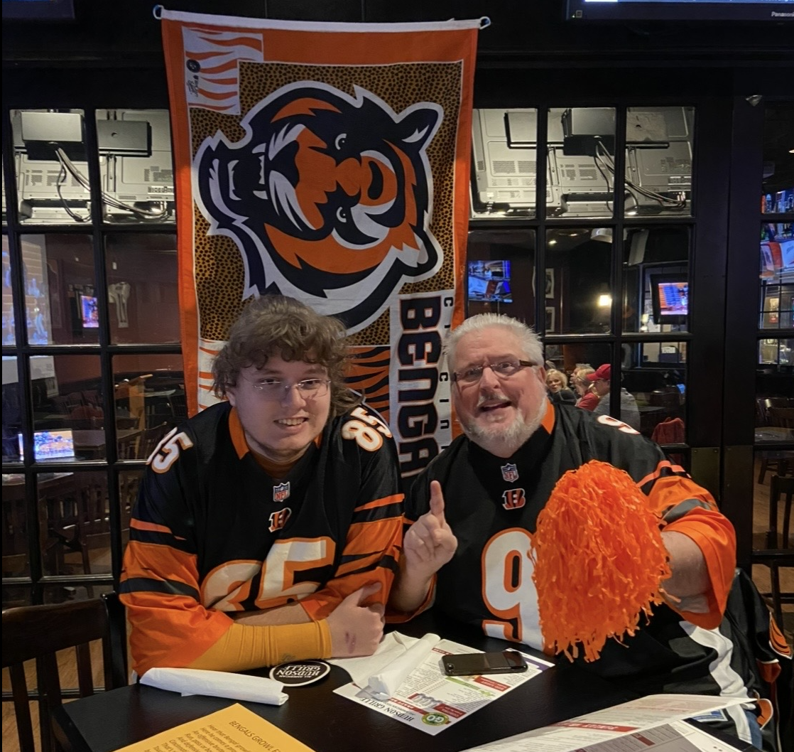 A father in son in Bengals jerseys smile and pose for a photo in a bar setting. There's a Bengals flag with a large stylized tiger and the word "Bengals" on it.
