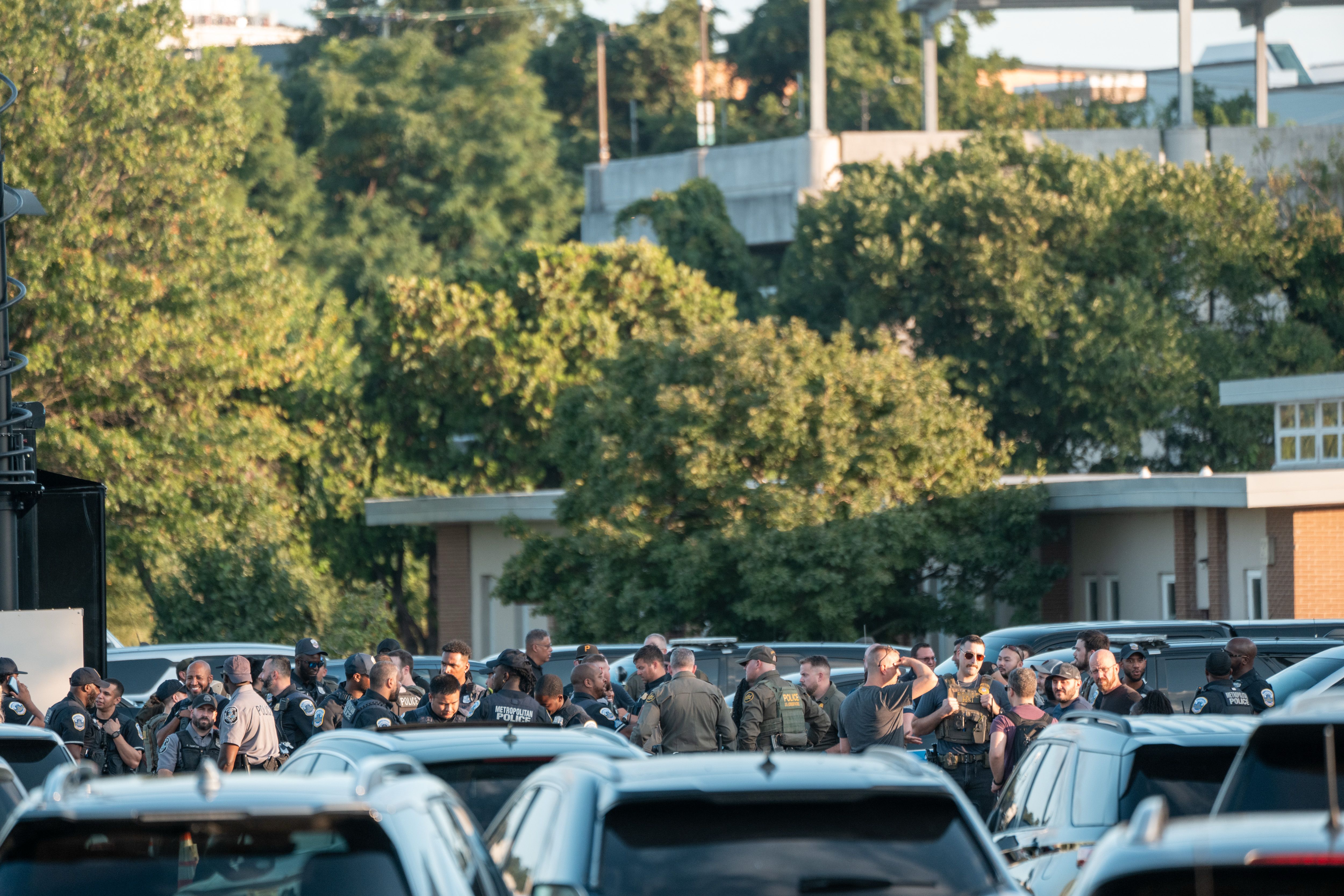 A large group of police officers in uniform gathered closely in a parking lot with many cars around, green trees and a building in the background during daylight.