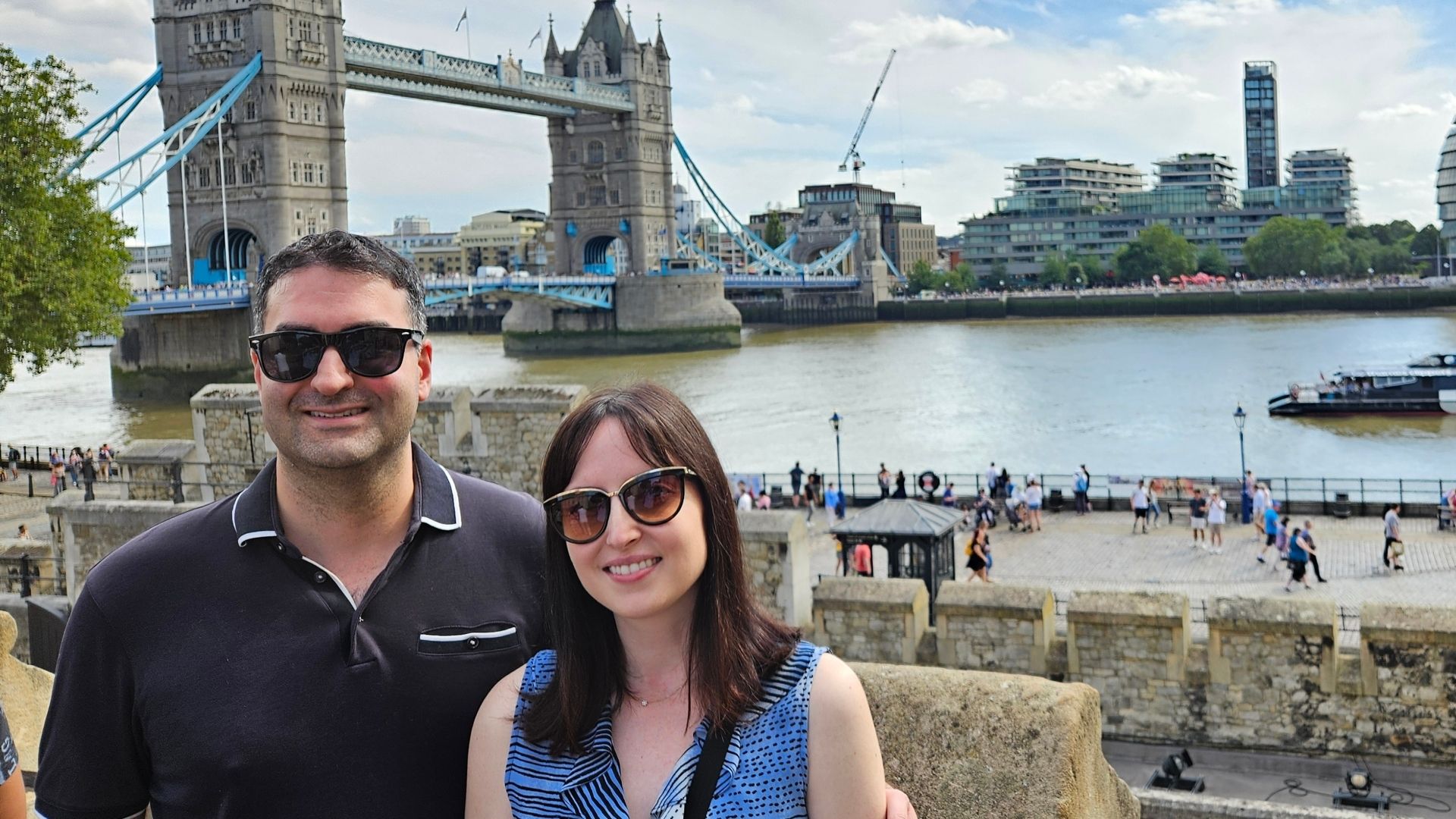 A smiling couple poses in front of the Tower Bridge in London, with the River Thames and modern buildings in the background.