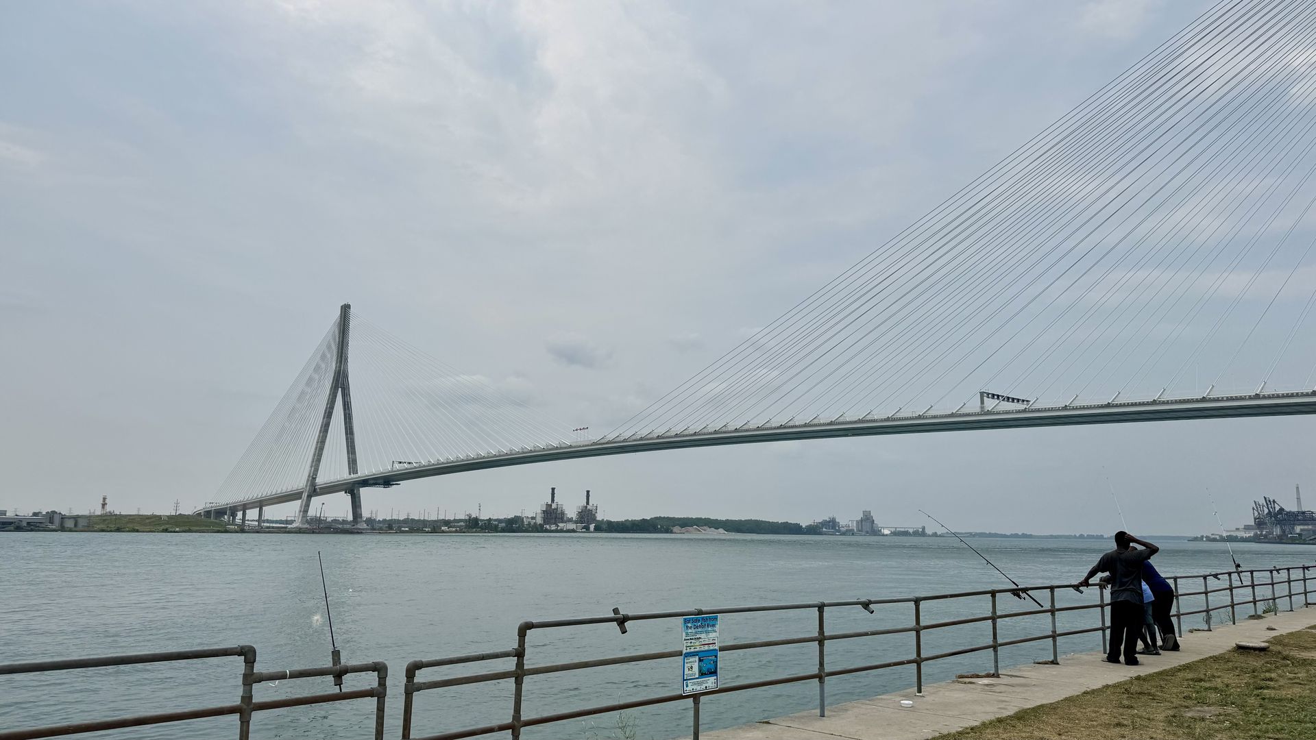 Cable-stayed bridge with a tall central pylon and many cables spans a wide river. Foreground shows a railing and two people by the water, with distant industrial buildings on the horizon.