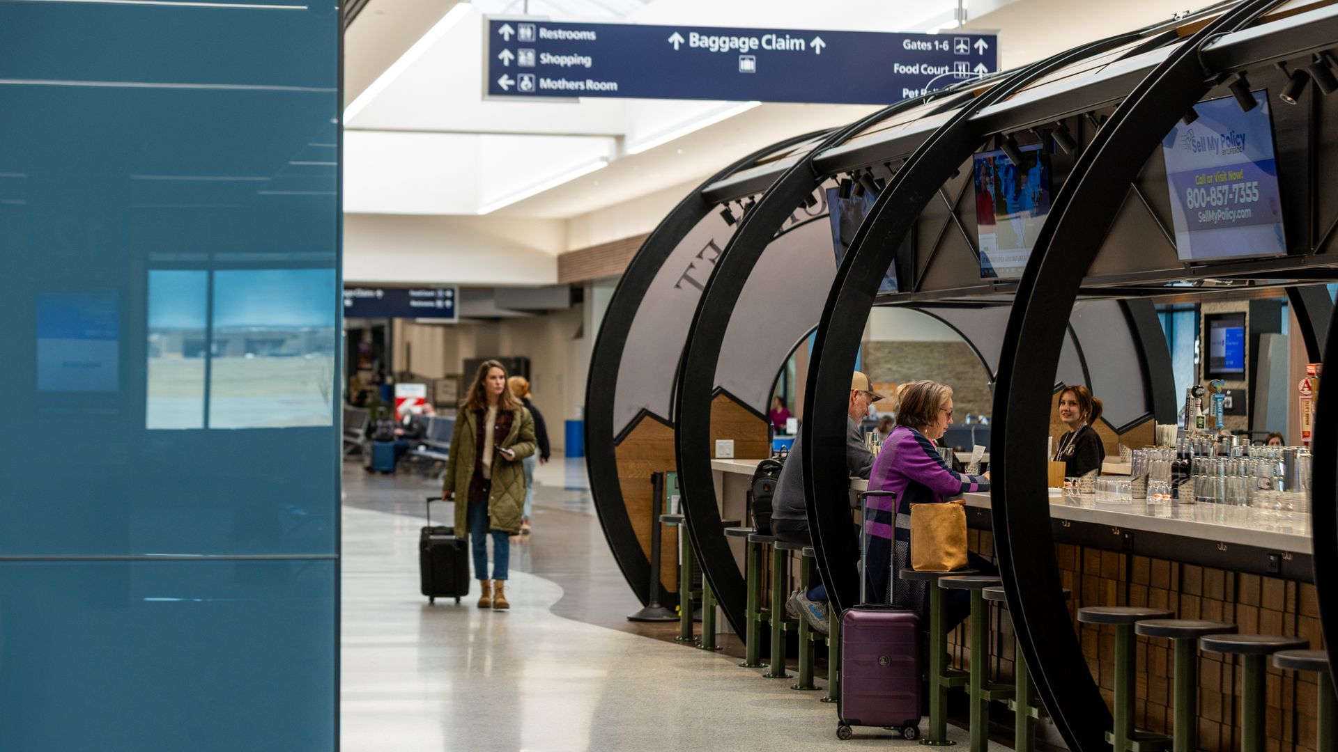 Inside an airport terminal, a blue wall on the left and curved black arches with stools on the right; people sit at a counter while an overhead sign points to Baggage Claim, Restrooms, and shops.