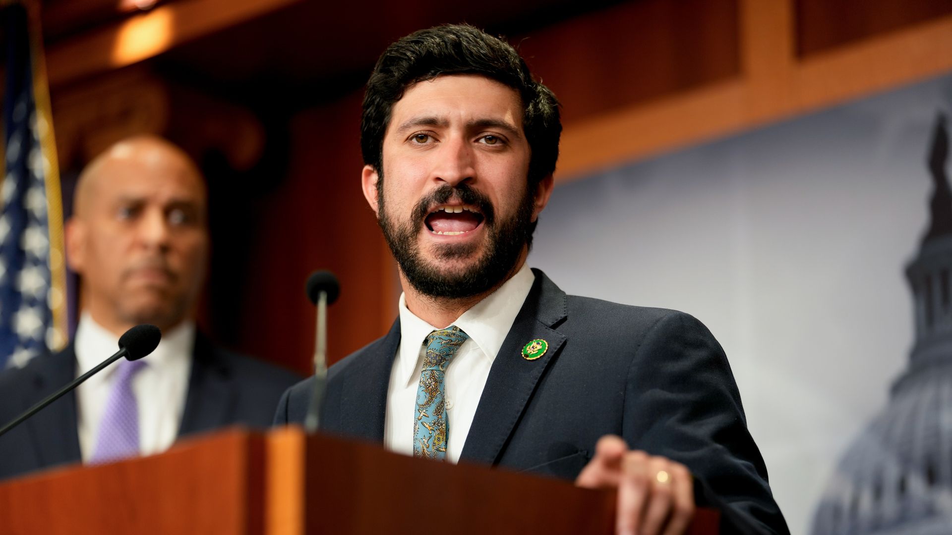 Rep. Greg Casar wearing a gray suit and speaking at a wooden podium.